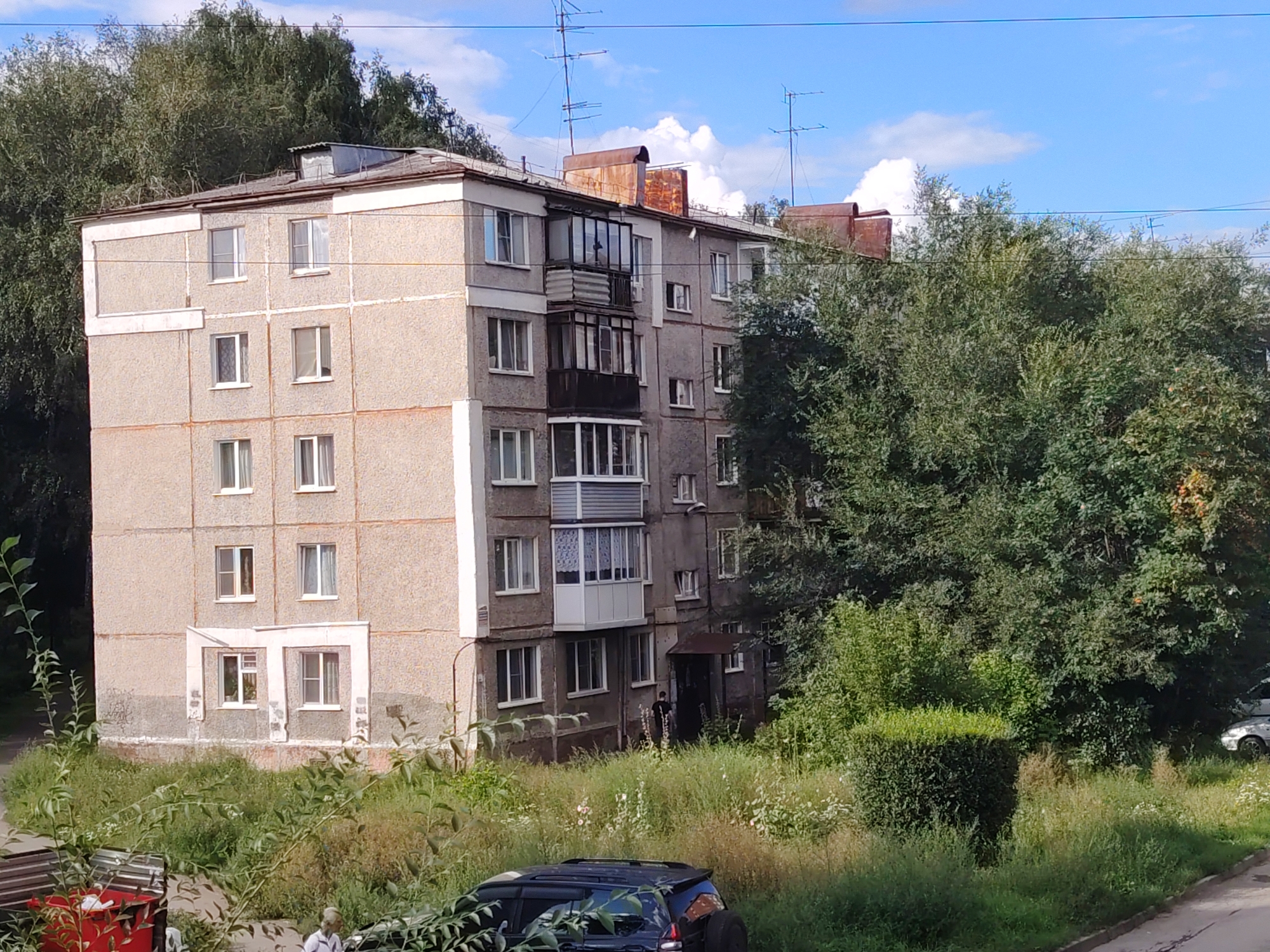 The image captures a daytime scene in Kemerovo, Russia, featuring a multi-story apartment building surrounded by overgrown vegetation. The building, appearing to be of Soviet-era construction, is a light-colored, concrete structure with multiple windows and balconies. The windows are mostly white-framed, and some balconies are enclosed with glass. Several television antennas are visible on the roof of the building.
The foreground and surrounding areas are characterized by tall, unkempt grass, bushes, and trees, indicating a lack of extensive landscaping. A dark-colored SUV is parked in the lower right portion of the image, partially obscured by foliage. To the left, a red recycling bin and part of a bench are visible. A person in a light-colored shirt and dark pants is standing near the entrance of the building, and another person in dark clothing is standing in the doorway of the building. The sky is partly cloudy with patches of blue. The overall impression is of a residential area with a somewhat neglected appearance.