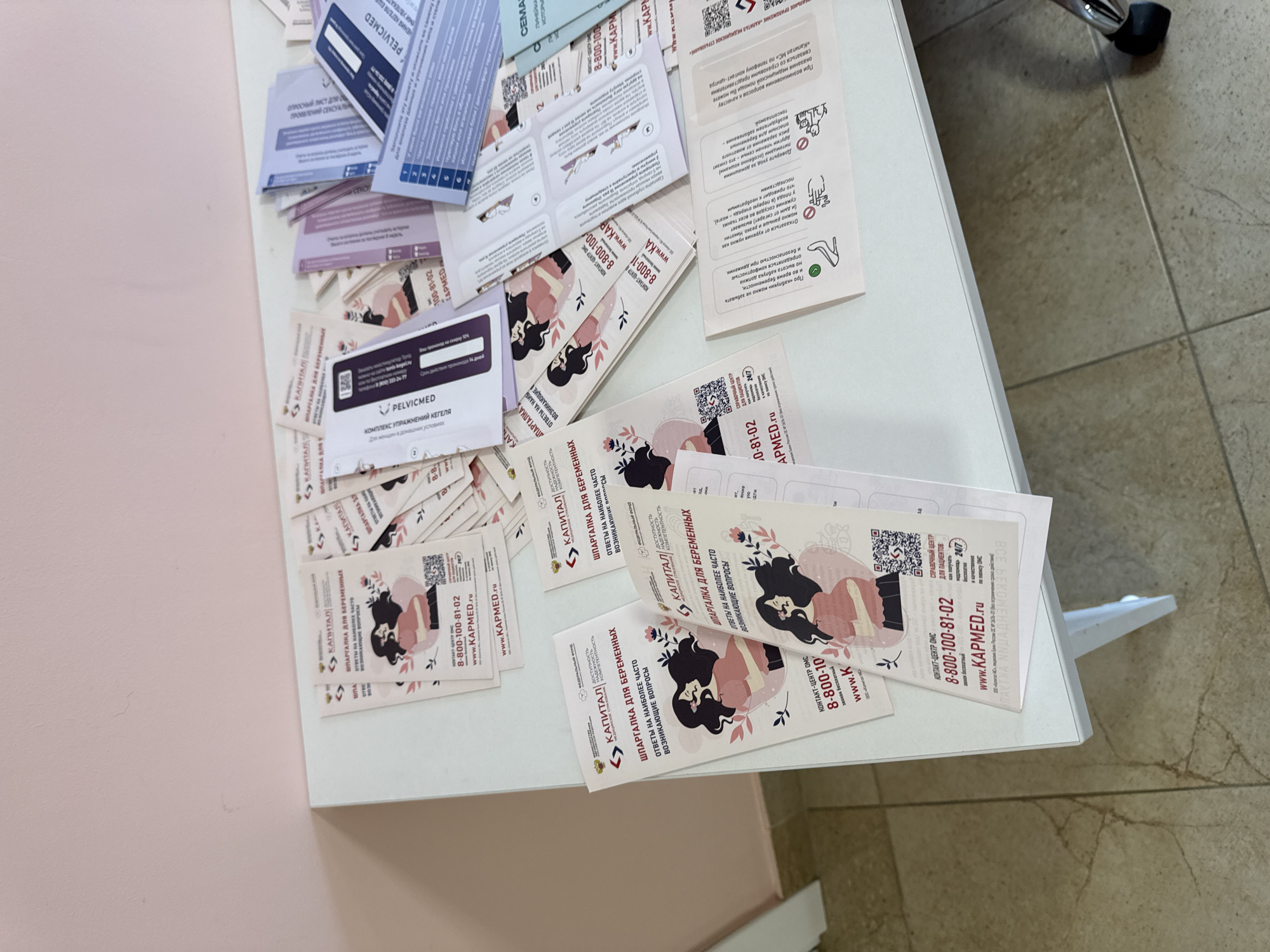 A white table in a room with pale pink walls is covered with numerous printed materials, mostly brochures and flyers. The brochures have a similar design featuring an illustration of a pregnant woman and text in Russian. Some of the text visible includes "ШПАРГАЛКА ДЛЯ БЕРЕМЕННЫХ" (Cheat sheet for pregnant women) and a phone number "8-800-100-81-02" along with a website "WWW.KAPMED.RU". There are also some other printed materials with different designs and text. A silver chair with a black base is partially visible on the right side of the image. The room appears to be well-lit, possibly from overhead lighting, casting shadows on the table and floor. The floor is tiled with a beige and brown pattern. There are no people visible in the image. The scene is consistent with an office or a waiting area where informational materials are distributed. The presence of specific Russian text and the context of pregnancy brochures suggest this location is likely in Russia. The city of Oktyabrskoye is mentioned as a possible location, which is a common place name in Russia.
