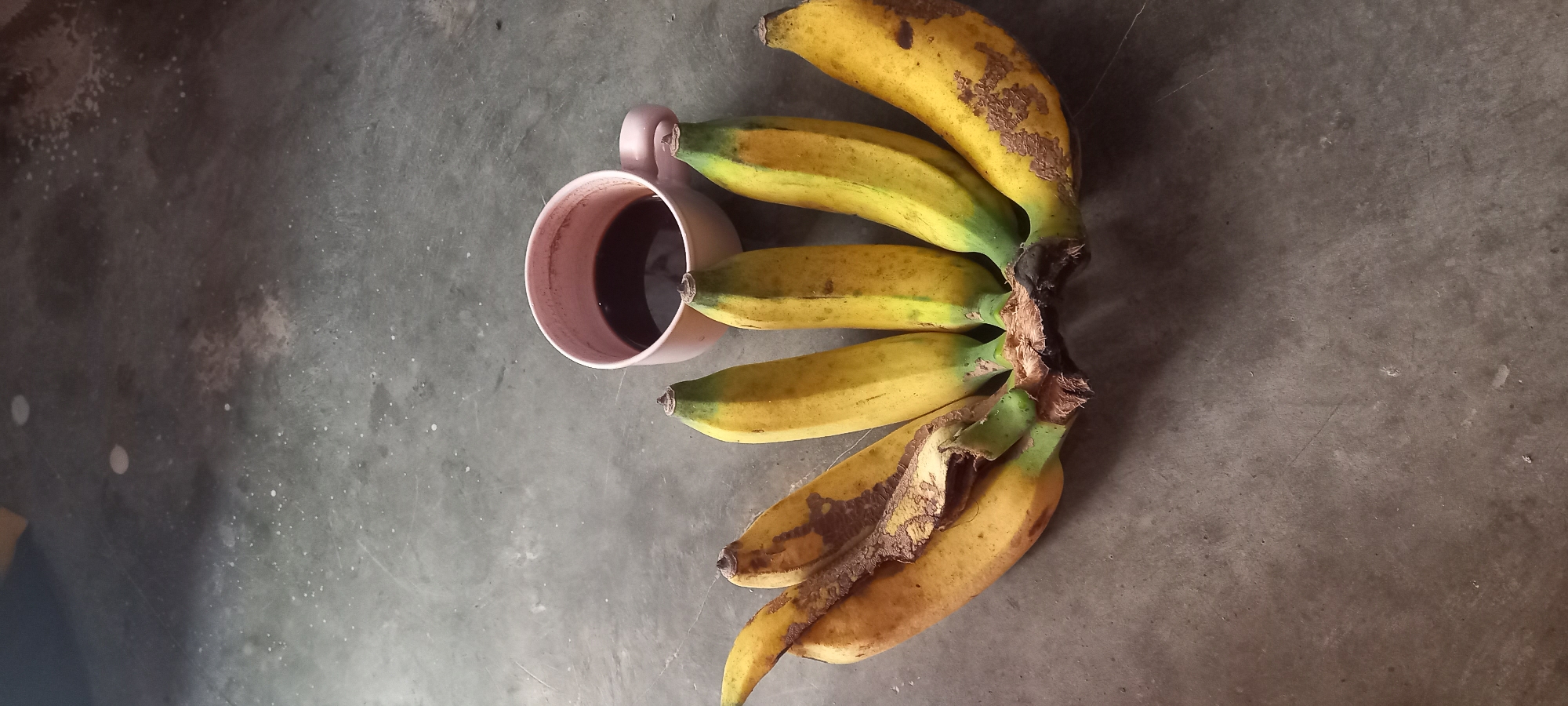 A bunch of six bananas and a light pink mug filled with a dark liquid are placed on a concrete floor in Sidamulya, Indonesia. The bananas are yellow with some brown spots, and the mug has a handle. The floor is a rough, gray surface with some lighter speckles and a few smudges. The lighting suggests an indoor setting.