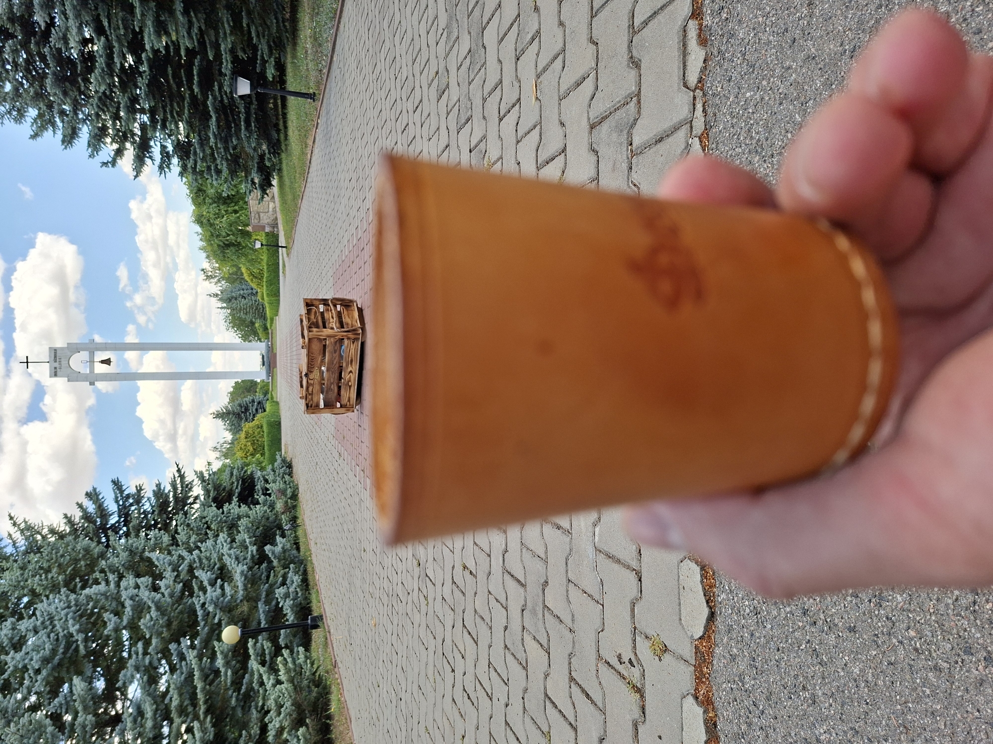 In the foreground, a person's hand holds a light brown, leather-like cup. The cup is slightly out of focus, with a visible stitched seam around its base. The background depicts a paved pathway leading towards a tall, white structure that resembles a bell tower, topped with a cross. The pathway is lined with tall, green evergreen trees. In the middle ground, on the pathway, sits an empty wooden crate. The sky is blue with scattered white clouds. The scene appears to be located in the city of Șerpeni, Moldova, likely a park or memorial area given the architectural elements and landscaping.