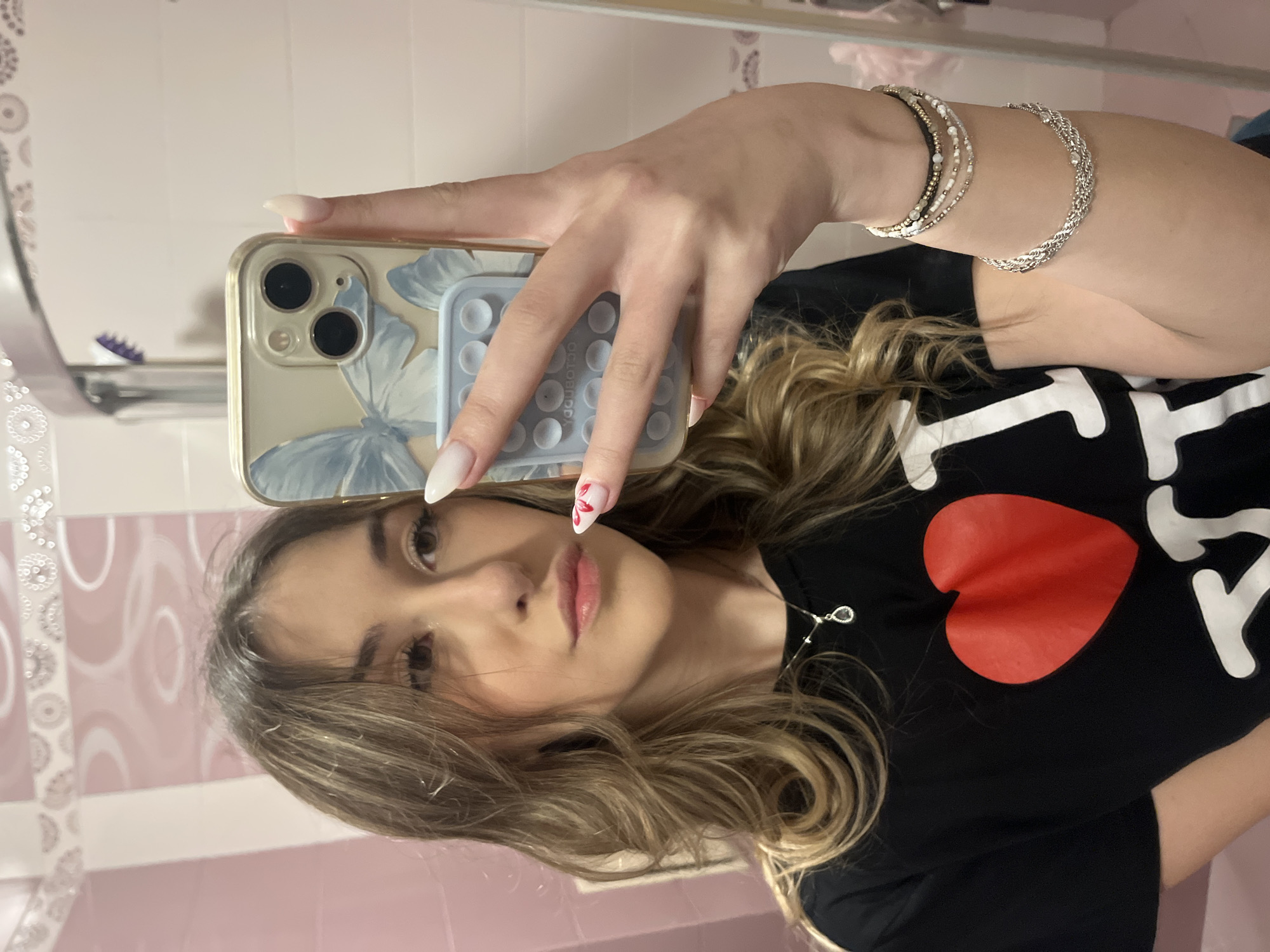 A young woman is taking a selfie in a bathroom. She is holding a phone with a butterfly-patterned case. The phone has a blue grip with indentations on the back. She is wearing a black T-shirt with a red heart on it. She has several bracelets on her arm. Her fingernails are long and have designs on them. The bathroom has pink and white patterned walls. The location is Sofia, Bulgaria.