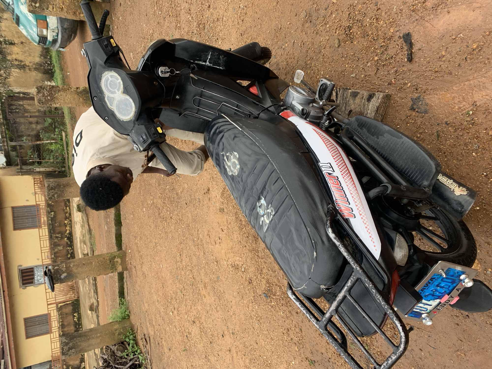 An overhead, medium shot shows a Black man wearing a light-colored, long-sleeved shirt and light-colored pants bending over a black scooter. The scooter is parked on a dirt road in front of a yellowish building. The man's face is obscured by his bent posture. The scooter has a black seat, black handlebars, and a black body. There is a license plate on the back of the scooter that is blue with white lettering and numbers. The building behind the man appears to be a house with windows and a red roof. The ground is covered in dirt and there are some scattered rocks and debris. The location is identified as Rafin Par, Nigeria.
