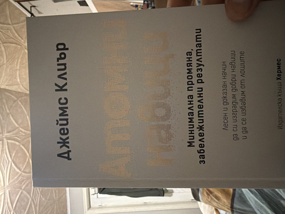 The scene depicts a paperback book held by a human hand, positioned prominently in the foreground. The book has a gray cover with text in Bulgarian. The author's name, "Джеймс Клиър" (James Clear), is printed in black at the top. The title, "АТОМНИ НАВИЦИ" (Atomic Habits), is rendered in a large, stippled, light orange-gold font below the author. Further text in black includes the subtitle "Минимална промяна, забележителни резултати" (Minimal change, remarkable results) and a descriptive phrase "Лесен и доказан начин да си изградим добри навици и да се избавим от лошите" (An easy and proven way to build good habits and get rid of bad ones). The publisher, "Издателска къща Хермес" (Hermes Publishing House), is indicated at the bottom. A light-toned human hand is partially visible on the right side, gripping the book. The background includes an ornate, light-colored patterned ceiling or wall in the upper left. To the left, a light-colored door frame or window frame is partially visible, with a dark garment hanging on a hook behind it. The lower left shows blurred textiles, possibly bedding, in warm tones. The lighting suggests an indoor setting. This scene is located in Drachevo, Bulgaria.