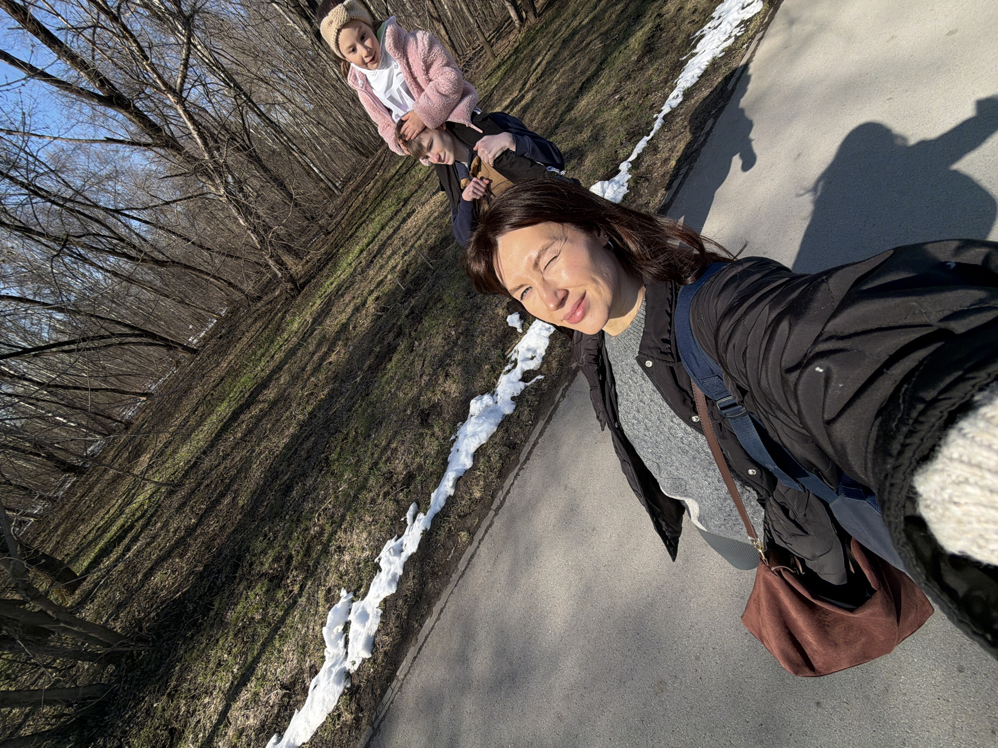 A selfie photograph taken at eye-level shows three people on a paved path next to a park setting in Perm, Russia. The foreground features a woman with dark hair, wearing a black jacket, grey sweater, and a brown shoulder bag. She is smiling. Behind her is a younger male subject. Behind the male subject is a young girl wearing a pink sweater and a beige headband. A patch of snow is visible along the edge of the path. Bare trees line the park area. The sky is clear. The path is paved. The photograph captures the subjects during the daytime.