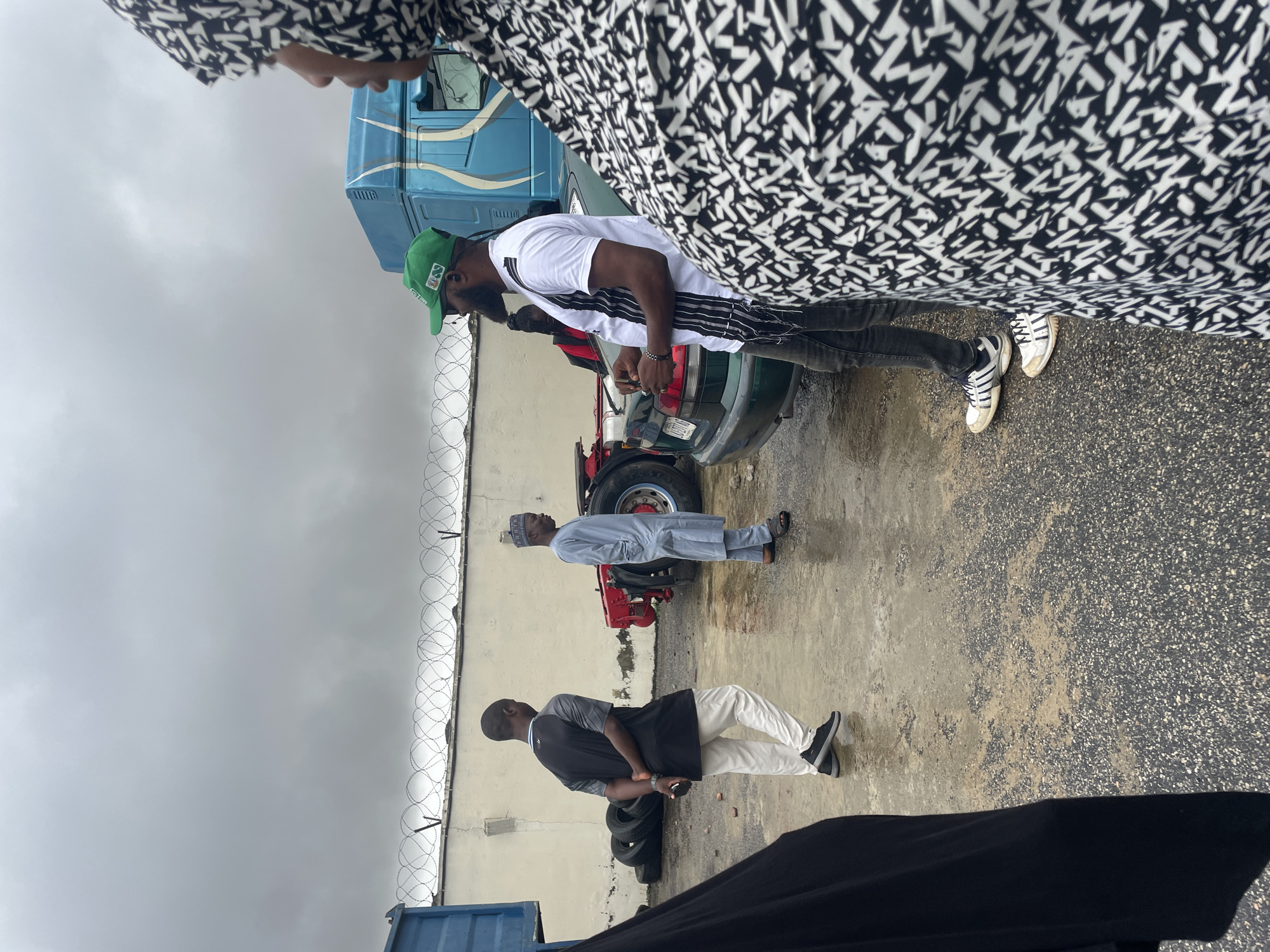 The image depicts a group of people outdoors in what appears to be a truck stop or industrial area, given the presence of a large blue truck and stacked tires. The ground is wet, suggesting recent rain. A coiled barbed wire fence runs along the side of a light-colored building, indicating a secure or potentially industrial location. The sky is overcast, suggesting a cloudy day, possibly early morning or late afternoon.
In the foreground, a person wearing a black garment with a distinctive white and black abstract pattern is partially visible. Next to them, a person wearing a white t-shirt, dark pants, and a green baseball cap with some emblem is standing and looking downwards. Another individual in the middle ground is wearing a light blue traditional outfit and a cap, also looking downwards. Further back, a person in a black and grey t-shirt and white pants stands with their arms clasped behind their back.
The people seem to be engaged in some form of waiting or observation, with their gazes directed downwards or slightly away. The overall mood is subdued, possibly due to the weather or the nature of the activity. There is no visible text in the image. The setting, with the truck, tires, and industrial fence, along with the overcast sky and wet ground, strongly suggests an outdoor, functional environment in Lokoja, Nigeria.