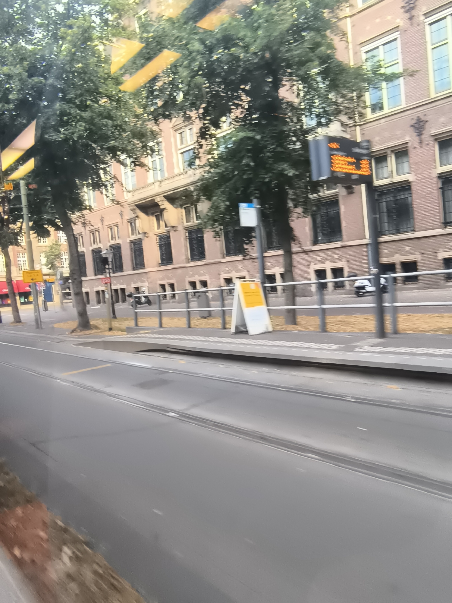 The image shows a street scene in The Hague, Netherlands, likely a tram stop. In the foreground, a grey asphalt road with tram tracks is visible. The background features a multi-story brick building with numerous windows. Trees with green foliage line the sidewalk.
Several objects are present:
- A digital display board with orange illuminated text, likely indicating public transport schedules, is mounted on a pole.
- A white A-frame sign with yellow text and a black arrow is placed on the sidewalk.
- A silver trash bin is located near a metal railing.
- A white scooter is parked behind the railing.
- A yellow directional sign with an arrow is visible on a pole further down the street.
The scene appears to be during daytime, with sunlight casting shadows. The overall impression is one of a typical urban street environment in The Hague.