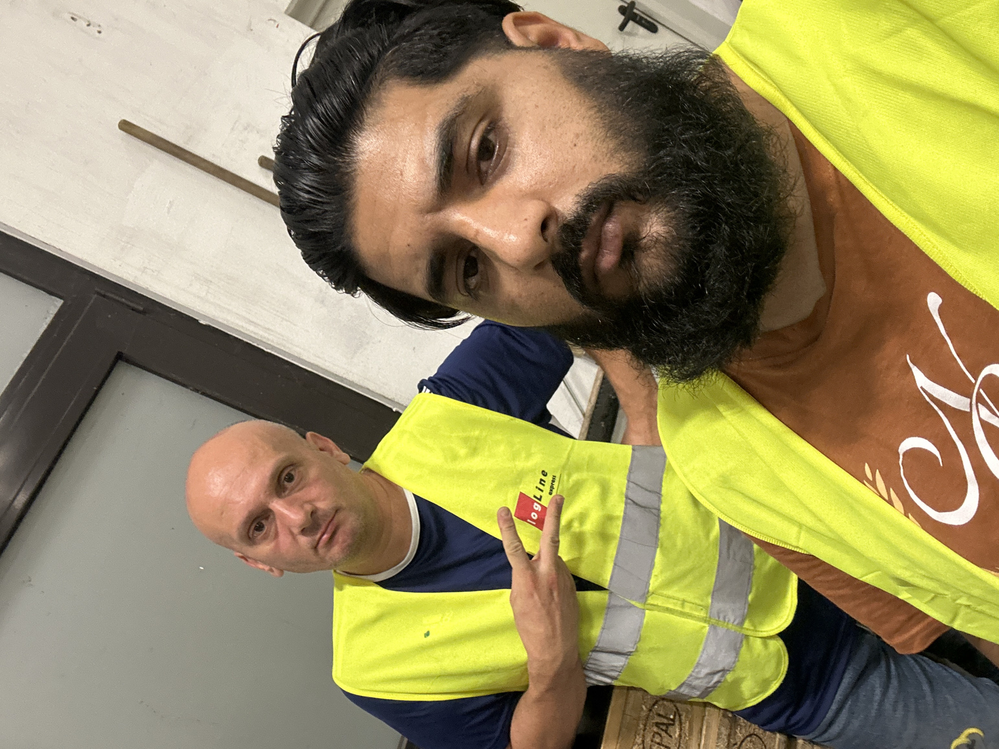 In a dimly lit indoor space, possibly a warehouse or industrial facility in Schweitenkirchen, Germany, two men are positioned for a selfie. The man on the left, bald with a pale complexion, wears a dark blue t-shirt and a bright yellow high-visibility vest with reflective stripes. He is looking directly at the camera with a neutral expression and is holding up two fingers in a peace sign. His vest displays a red logo with white text that appears to read "logLine express".
The man on the right has dark hair slicked back, a prominent beard, and is also wearing a yellow high-visibility vest over a brown t-shirt with white text that appears to be lettering. He is closer to the camera and his gaze is directed slightly off-center. The background consists of a light-colored wall, a dark door frame or window, and stacked wooden pallets to the left, one of which has "PAL" printed on it. The overall lighting suggests it may be evening or an interior with artificial light.