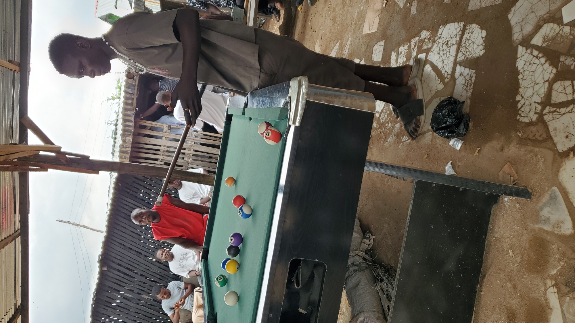 In Jalingo, Nigeria, a group of men gather around a green billiard table outdoors, beneath a rustic, open-sided structure with a corrugated metal roof. A man in a gray t-shirt and dark shorts stands to the right, holding a billiard cue, poised to take a shot. He wears a white beaded necklace. Scattered on the table are several billiard balls, including a white cue ball, a red and white striped 11-ball, a solid blue 7-ball, a solid purple 5-ball, a solid black 3-ball, a solid yellow 1-ball, a red and white striped 15-ball, and an orange 9-ball. In the background, other men are seated or standing, observing the game. The ground is a mix of dirt and cracked concrete, with a black plastic bag near the feet of the man with the cue. The overall atmosphere suggests a casual gathering and recreational activity.