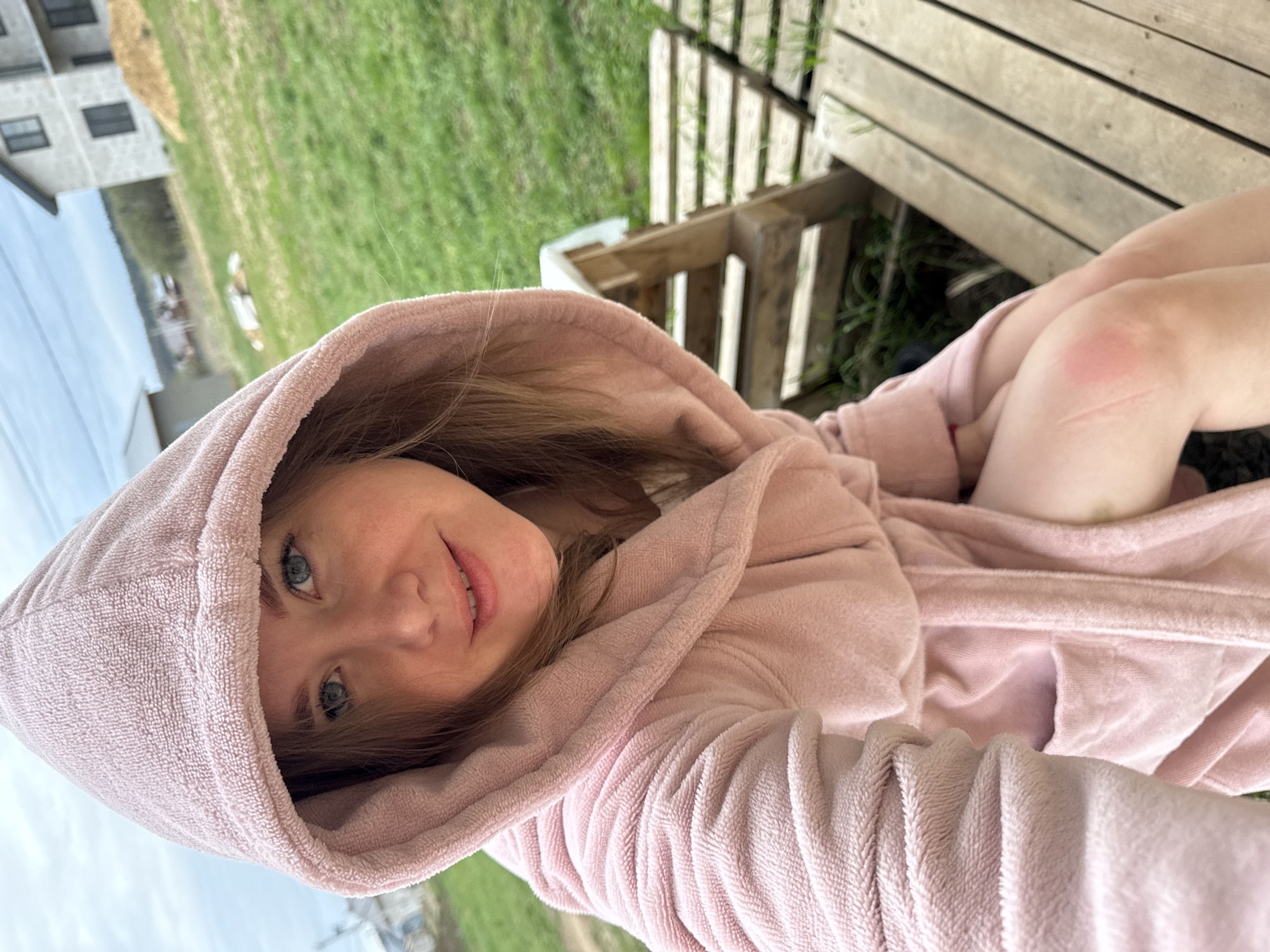 A young fair-skinned woman wearing a pink hooded bathrobe is outdoors. The woman is facing the camera, holding the camera at an angle so that the grass, part of a building, and a wooden structure are visible in the background. The location may be a backyard or porch area in the city of Ферма, Russia. A light bruise is visible on the woman's knee.