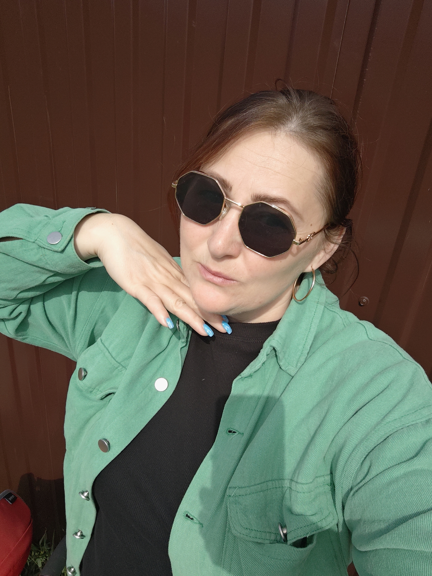 A woman poses for a portrait outdoors in Марковский, Russia, against a backdrop of a tall, dark brown corrugated metal fence. The scene is illuminated by bright, sunny daylight, creating clear highlights and shadows.
The woman is wearing large, hexagonal-framed dark sunglasses with gold accents, and golden hoop earrings. Her dark brown hair, styled back, shows some lighter roots near her forehead. She is dressed in a vibrant mint green, button-up jacket, likely made of denim or corduroy, worn over a black ribbed top. The jacket features prominent silver-toned snap buttons down the front and on the cuffs.
She holds her right hand gracefully to her neck and chin, displaying bright blue nail polish with a subtle patterned design. Her expression, largely obscured by the sunglasses, appears confident and self-assured, with a slight pout on her lips. In the lower-left corner of the frame, a hint of a red object, possibly a bag or a piece of furniture, is visible, along with some green vegetation at the base of the fence. There is no visible text in the image.