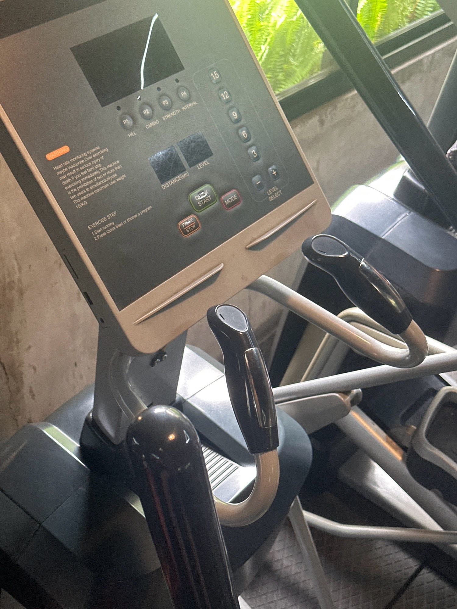 This image captures a close-up view of the console and handles of an elliptical exercise machine. The console features a digital display screen, various buttons labeled with functions like "HILL," "CARDIO," "STRENGTH," "INTERVAL," "LEVEL SELECT," "QUICK START," "MODE," and "PAUSE/STOP." It also shows numerical buttons ranging from 3 to 15, likely representing levels or programs. A warning label regarding heart rate monitoring systems is visible on the left side of the console. The handles of the elliptical are made of black rubber grips with silver accents, attached to gray metal arms. The background includes a glimpse of green foliage through a window and a concrete wall. The scene is indoors, with the elliptical machine appearing to be in a fitness facility. The image does not provide any information to place it specifically in Lalitpur, Nepal, or any other city.