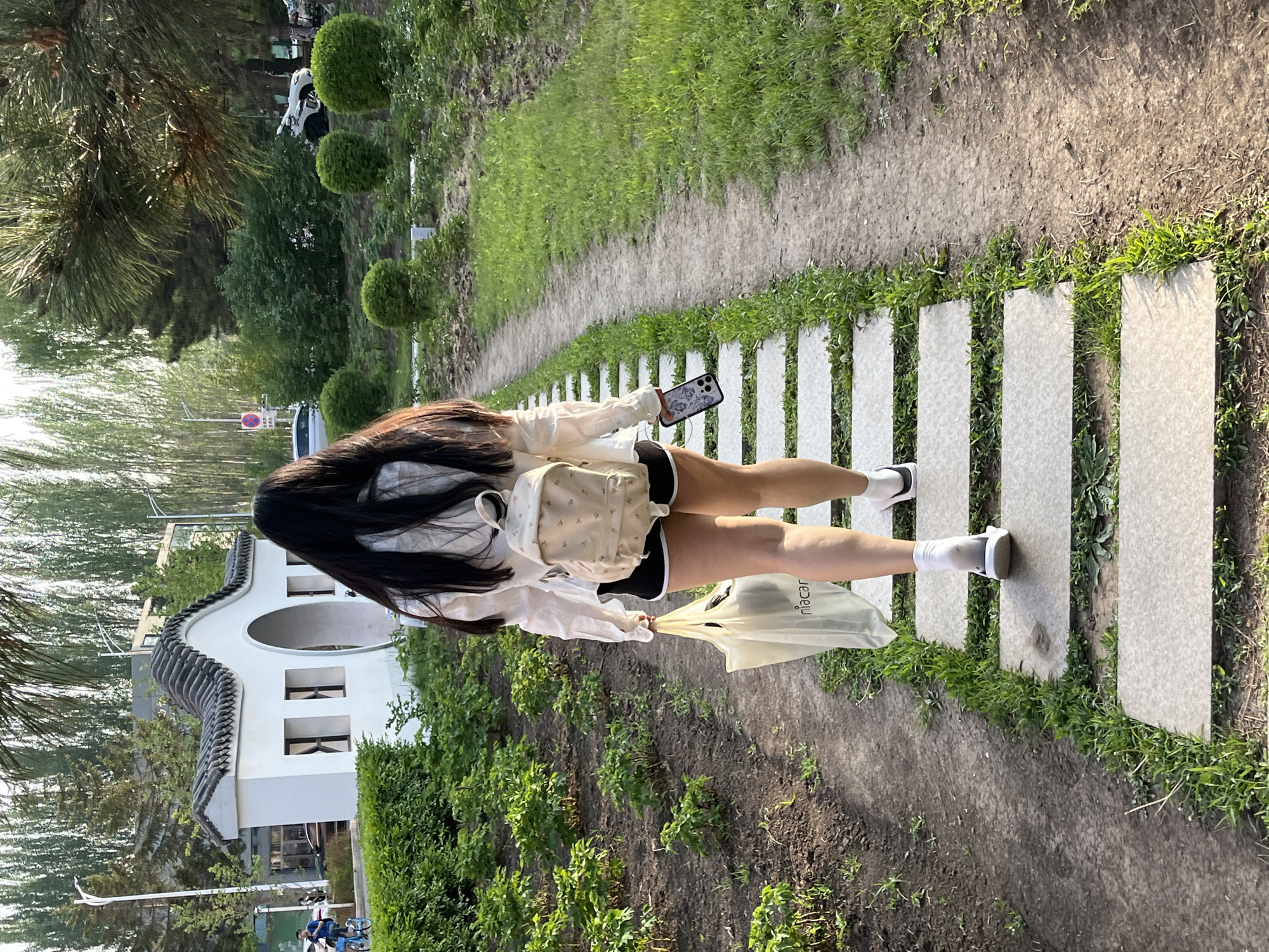 A waist-down shot shows a young woman walking on a path made of rectangular stones in Changchun City, China. She has long, dark hair and is wearing a white long-sleeved shirt, black shorts, white socks, and white sneakers. A small beige backpack is slung over her shoulder. She holds a smartphone in her right hand and a white plastic bag with the word "niacar" printed on it in her left hand. The path runs through a grassy area with various plants. In the background, there is a traditional Chinese building with white walls and a gray tiled roof, as well as trees and other greenery. A paved road and other buildings can be seen in the distance.