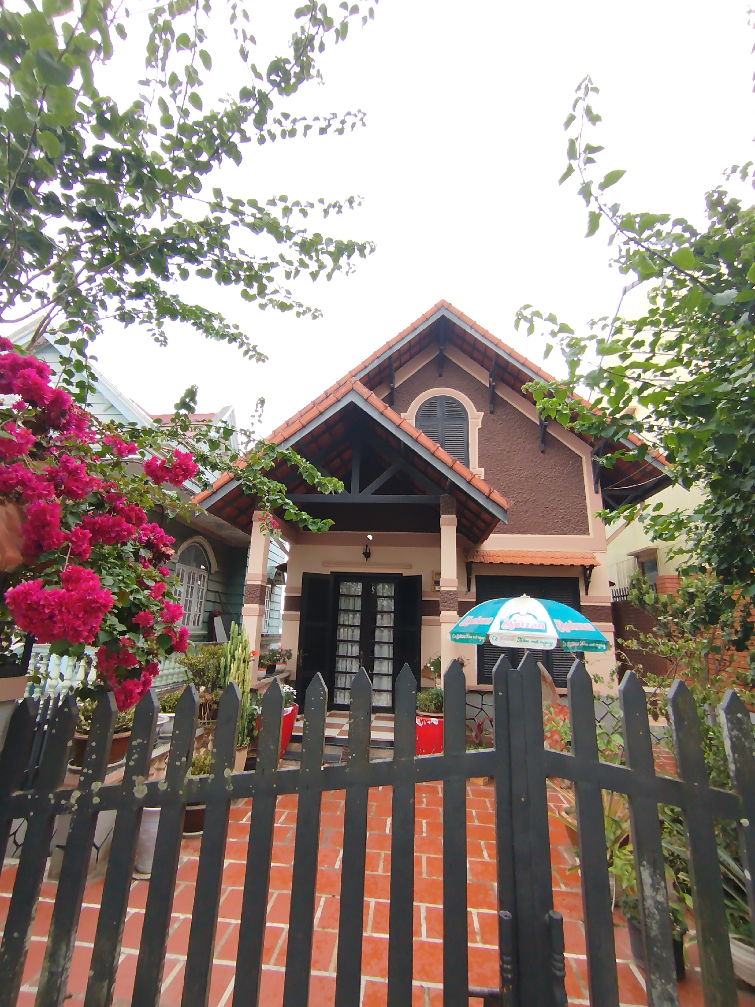 A ground-level, outdoor shot shows a house in Lâm Viên, Đà Lạt, Vietnam. The house has a brown and beige exterior, with a tiled red roof. The front of the house features a black door with multiple small square glass panes, and a small awning above the entrance. A blue and white umbrella stands to the right of the door. In front of the house is a black wooden fence with vertical planks. Red bougainvillea flowers are on the left side of the frame. The ground in front of the house and behind the fence is tiled with orange brick-like pavers. Green trees and foliage surround the house, with branches extending into the top and sides of the frame. The sky is white and overcast.
