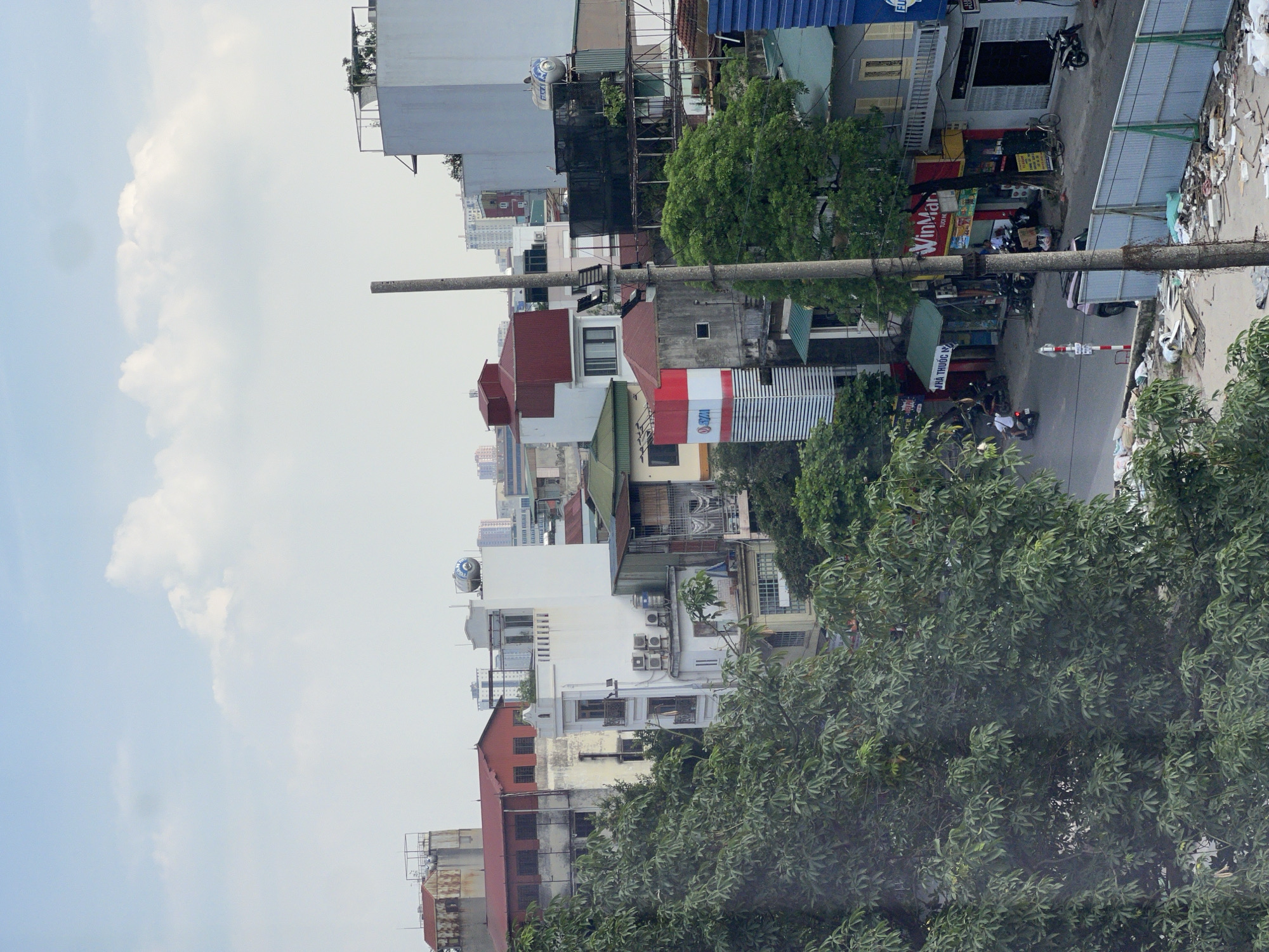This is a medium-shot, eye-level, outdoor, daytime, vertical-format photograph taken from a high vantage point, looking down and across a street in Hà Nội, Vietnam. The scene is predominantly urban, with a mix of buildings and trees.
In the foreground, a dense canopy of dark green foliage from a large tree dominates the left side of the frame, partially obscuring the view of the street and buildings below. A concrete utility pole stands prominently in the mid-ground, slightly off-center to the right, with its rough texture visible.
The street below is a mix of asphalt and some debris, particularly on the right side where construction materials and trash are piled near a pale blue metal fence. Several parked motorcycles and bicycles are visible on the street. A few people are present: one person rides a white scooter, another person is seated behind a stall, and a few other figures are obscured by distance and foliage.
A variety of buildings line the street. On the left, a multi-story white building with balconies and air conditioning units is visible. To its right, a building with a red roof and weathered facade stands. Further down the street, a building with a prominent red and white striped facade featuring the "WinMart" logo is a key feature. Other buildings show varied architectural styles and colors, with signs indicating businesses and residential spaces. In the background, taller urban structures and a water tower on a rooftop are visible against a cloudy sky.
The overall impression is a bustling urban environment, characterized by dense construction, green spaces, and everyday street life.