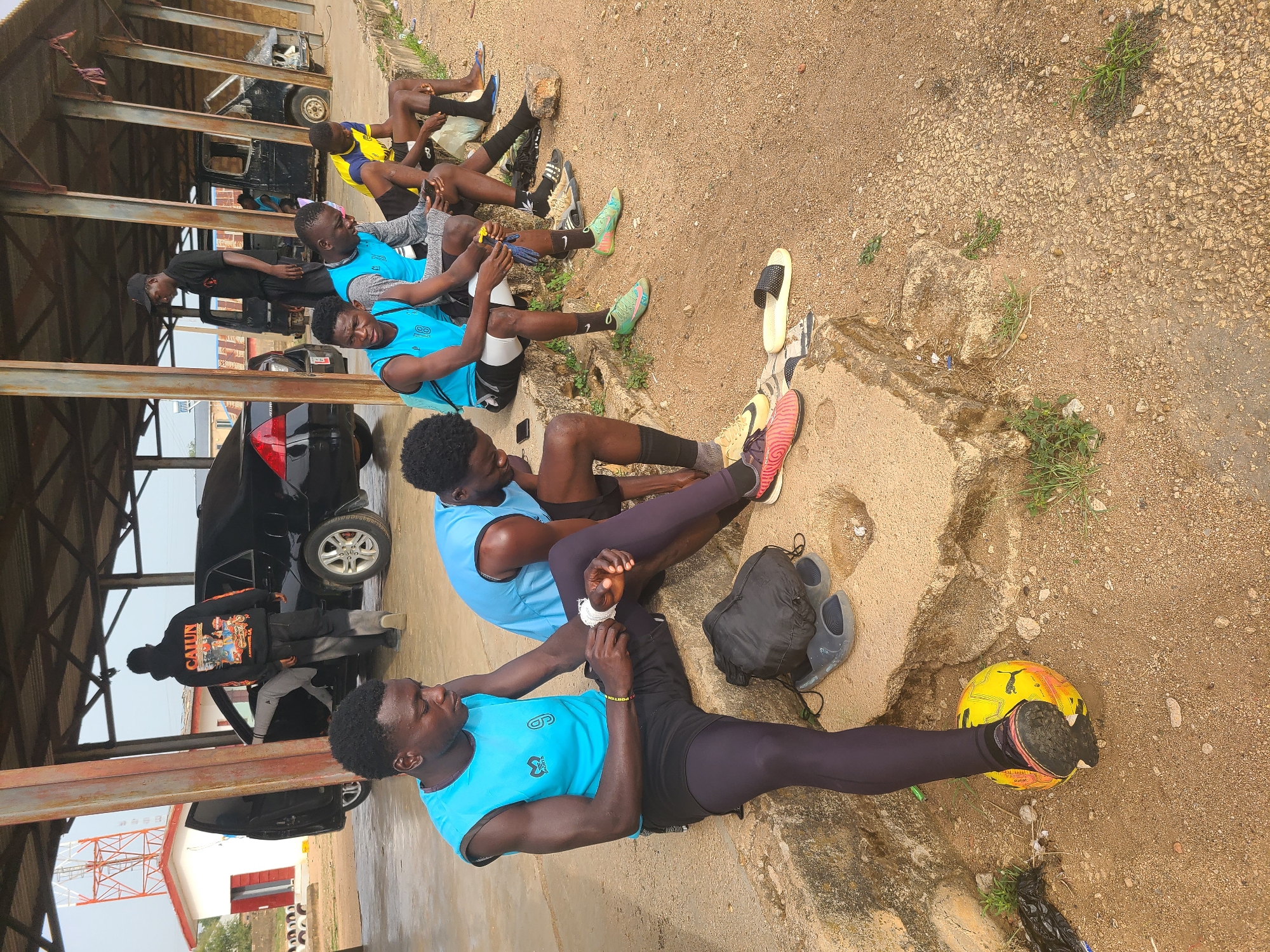 The image shows a group of six young men resting outdoors, seemingly after a physical activity. They are sitting on a low concrete structure along the edge of a dirt and gravel ground.
Four of the men are wearing similar light blue sleeveless shirts and dark-colored shorts or leggings. One man is wearing a yellow and blue shirt, and another is wearing a black sweatshirt. They appear to be stretching, tending to their feet, or simply relaxing. One of the men is wrapping a bandage around his wrist.
A yellow and orange soccer ball is visible next to one of the men. There are also sandals and a black bag on the ground.
Behind the men, there is a metal structure with a roof, under which several parked cars are visible. In the background, there is a red and white telecommunications tower, a building, and a glimpse of the sky.
The location is in Jos, Nigeria.