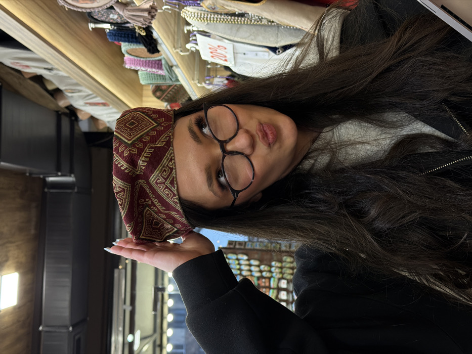 A single young adult female is depicted from the mid-chest upwards, observed inside a souvenir or hat shop in Yerevan, Armenia. She is trying on a maroon beret-style hat adorned with gold geometric patterns. Her right hand, exhibiting light-colored manicured nails, rests on the hat's brim. She possesses long, dark brown, wavy hair and wears dark-framed eyeglasses. Her facial expression is characterized by pursed lips in a pout and an upward-right gaze. Her attire consists of a dark jacket over a light-colored hooded top. The background on the right reveals multiple wooden shelves displaying various hats and textile merchandise, including a white sign with "20%" printed in red. To the left, shelves with smaller decorative objects are visible. The shop interior features wooden paneling on the ceiling and walls, along with exposed metallic ventilation ducts and artificial lighting.