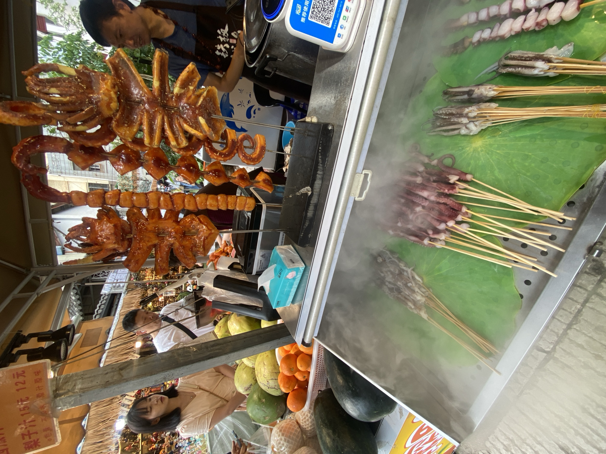 The image captures a vibrant street food stall, likely in Kunming, China, given the presence of Chinese characters and the style of the stall. The foreground is dominated by a stainless steel display case filled with skewers of seafood, predominantly squid and octopus, arranged on large green lotus leaves. Steam rises from the display, suggesting they are being kept warm or cooked. Behind the display, a variety of fruits, including watermelons, cantaloupes, and oranges, are piled up. A woman in a light-colored top is visible on the left, looking at the offerings. To her right, a man wearing a white t-shirt stands behind the stall, attending to customers or preparing food. Further back, another man, wearing an apron, is seen near a large pot and a payment terminal displaying a QR code, indicating modern payment methods are available. The stall is covered by a thatched roof, typical of many street markets. The overall scene depicts a bustling food market environment.
