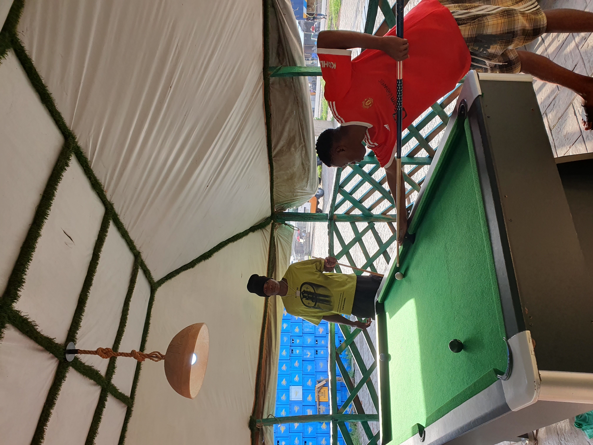In Lokoja, Nigeria, two young men are engaged in a game of billiards. The scene is outdoors, under a white tent supported by green wooden beams. The tent ceiling is decorated with artificial turf along the edges of the panels, and a light fixture hangs from the center. The billiards table is green with a dark frame and white corner protectors. One man, wearing a lime green t-shirt and a black beanie, stands near the table, holding a pool cue. The other man, dressed in a red Manchester United jersey and checkered shorts, is actively playing, leaning over the table with his cue. In the background, blue crates are stacked behind a green lattice fence, and a few people and vehicles can be seen in the distance.