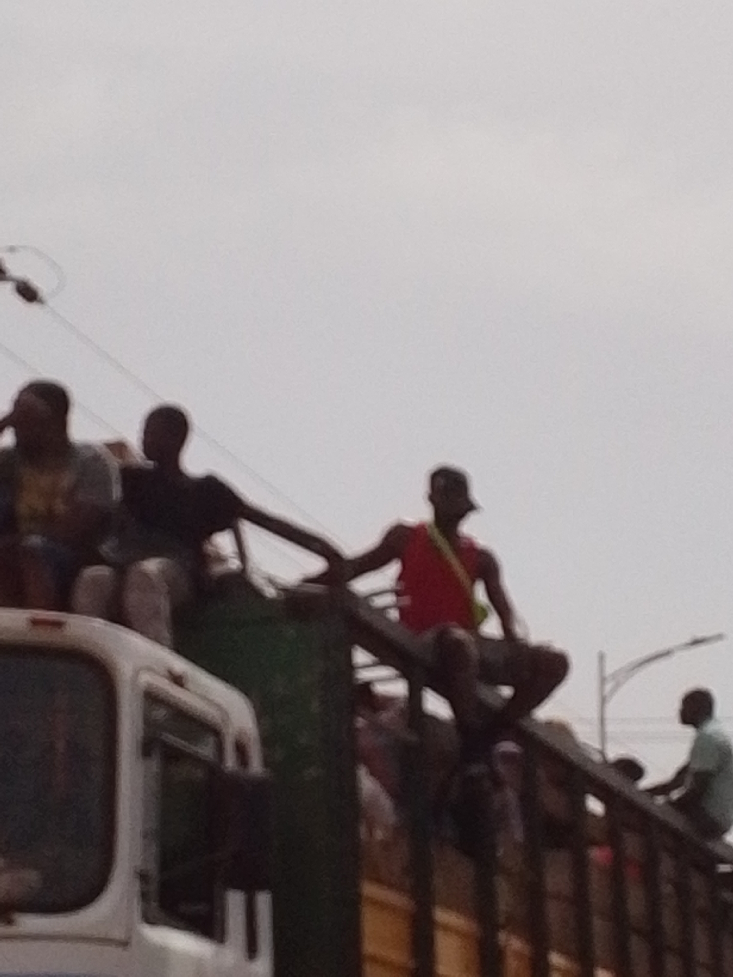 The image depicts several people riding on top of a large truck. The truck is white and green. The individuals are sitting and squatting, some are wearing casual clothes. The setting appears to be an urban environment, potentially the city of Tamale, Ghana. Overhead power lines are visible in the sky.