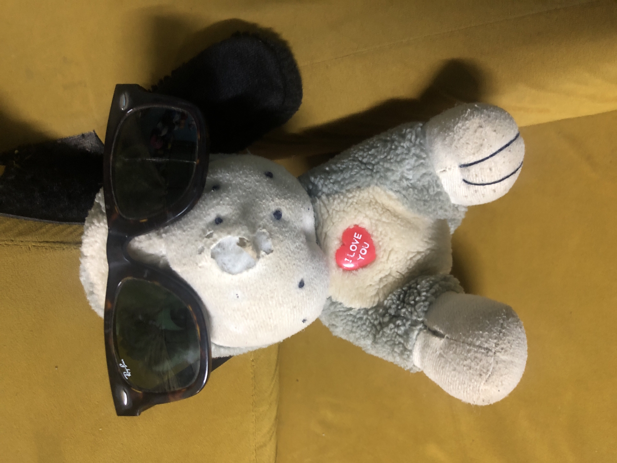 A stuffed toy dog with a heart-shaped button that reads "I LOVE YOU" sits on a yellow surface, possibly a couch. The dog is wearing sunglasses. The background is a textured yellow surface. The image is lit from above. There are no people visible in the image. The scene is indoors. The exact location in Lagos, Nigeria, is not discernible from the image.