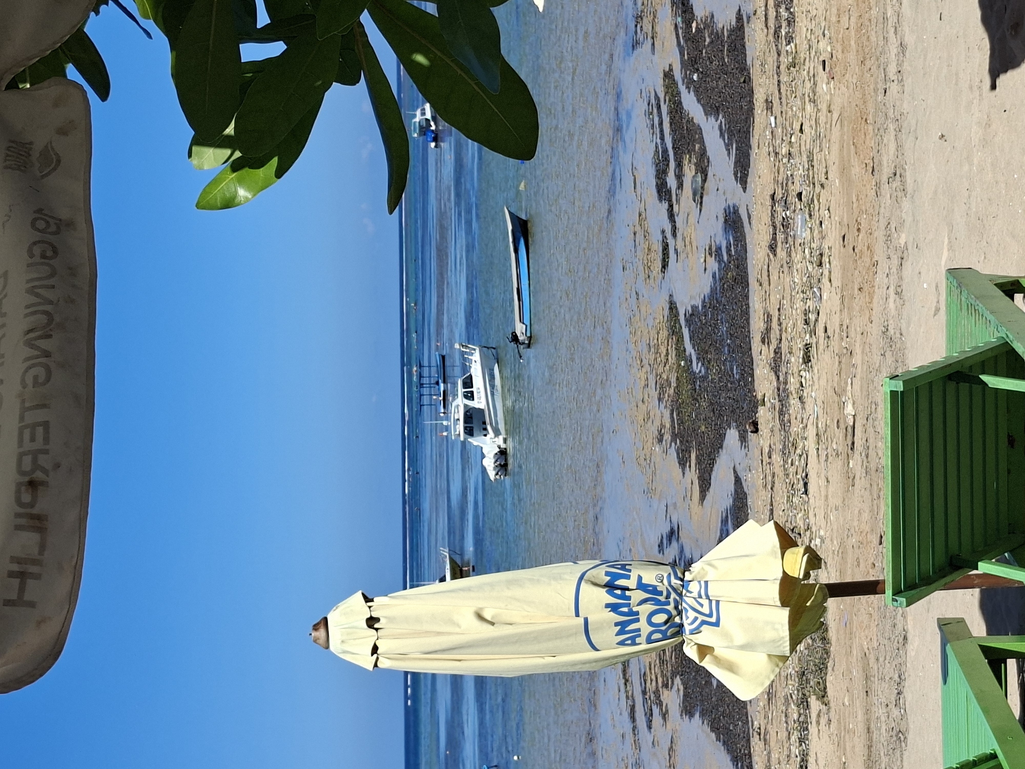 The image captures a beach scene near Denpasar, Indonesia. The vantage point is from behind a closed beach umbrella, with a green beach chair visible on the right. In the background, a calm, shallow bay is visible, with several boats anchored in the water. The beach is littered with seaweed and other debris. The sky is a clear blue, and some foliage is visible at the top of the frame. The brand name "Anantara Bora" is printed on the side of the umbrella.