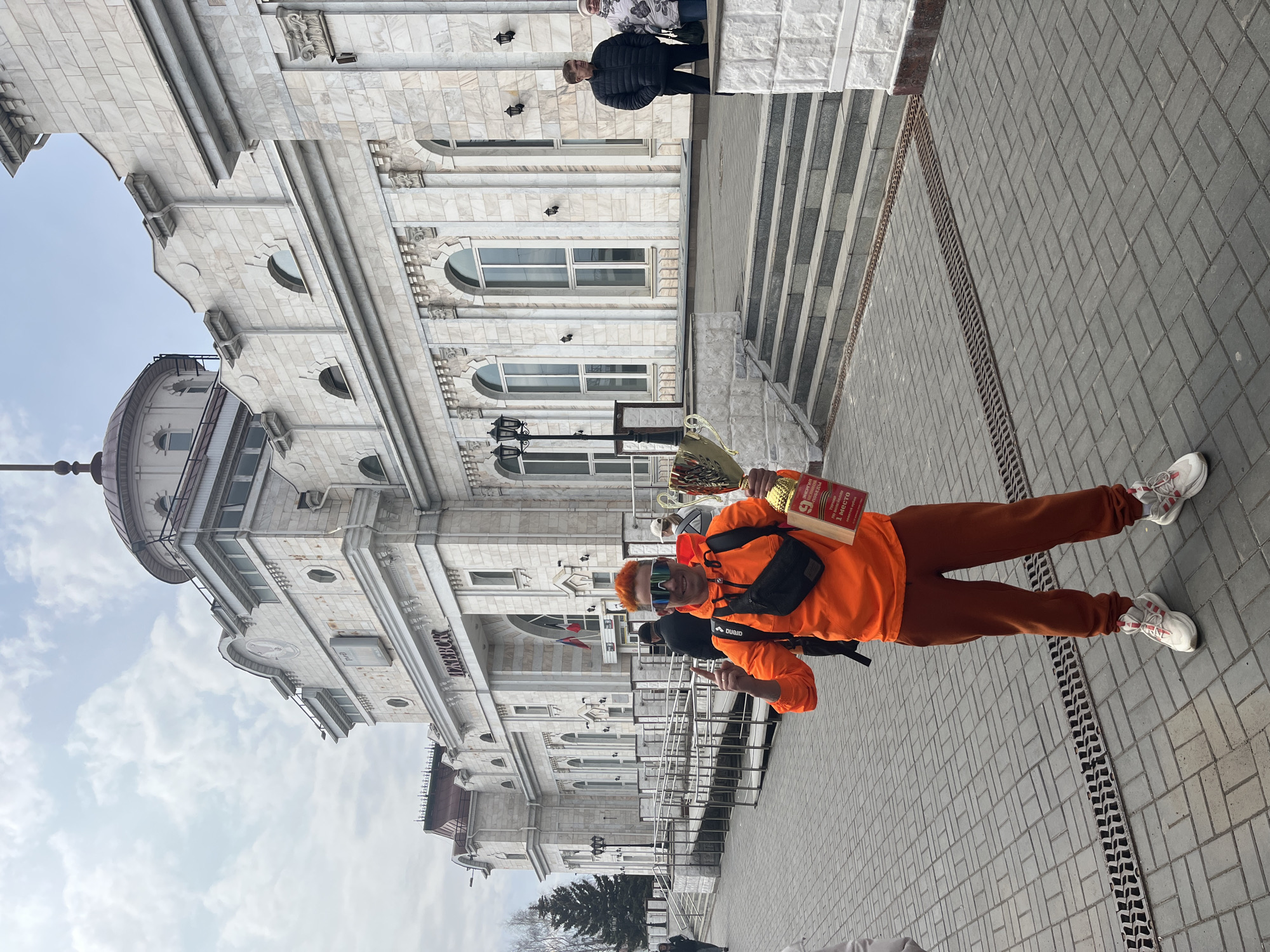 A man stands in front of the Izhevsk circus building in Izhevsk, Russia, holding a trophy and a book. He is wearing an orange sweatshirt, brown pants, and white sneakers with red stripes. The circus building is a large, white stone structure with multiple windows and a dome. The man is standing on a gray brick walkway. Another person is partially visible at the top of the stairs of the building.