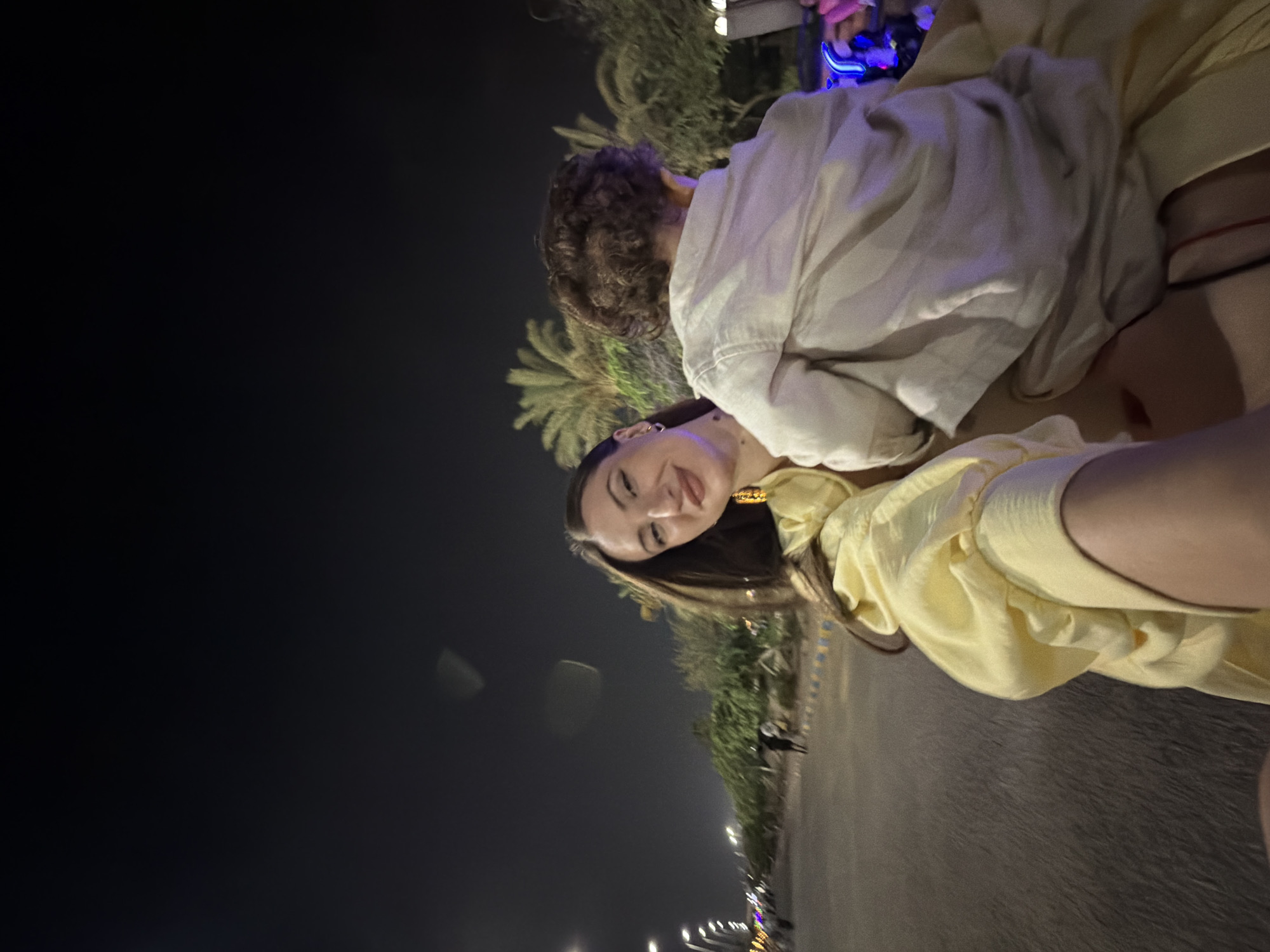 In a nighttime outdoor scene in the city of 6th of October, Egypt, a woman with long brown hair is holding a young child. The woman is wearing a light yellow top with voluminous sleeves and large, gold hoop earrings. The child, seen from behind, has curly brown hair and is wearing a light-colored shirt. They appear to be in motion, possibly on a vehicle, with palm trees and streetlights visible in the background. The road surface is visible in the foreground, with some blurred figures and vehicles in the distance. The sky is dark, indicating it is night.