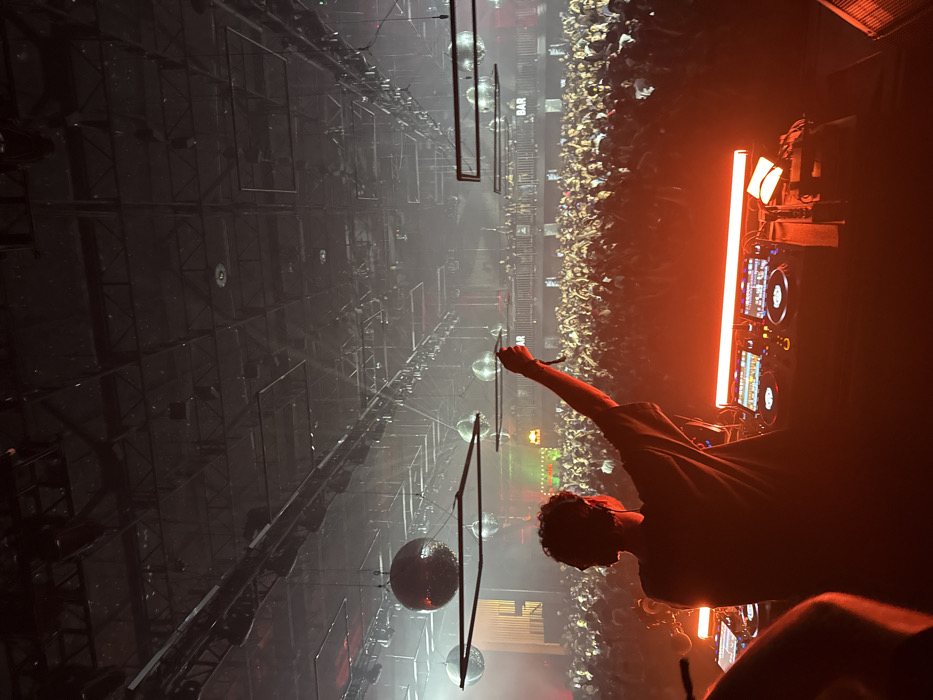 The image depicts a crowded indoor music venue, specifically identified as the Shrine Expo Hall in Los Angeles, California, United States of America.
In the foreground, a male individual, likely a disc jockey, is silhouetted from behind, facing a large audience. He wears a dark, loose-fitting shirt and has curly hair. His right arm is raised, holding a dark cable or lanyard. Before him rests a professional DJ setup, including two Pioneer CDJ-style decks and a mixer, all illuminated by a prominent orange-red horizontal light bar on the console. A bag or case is situated to the right of the equipment.
Beyond the DJ, a vast, densely packed crowd occupies the main floor, extending towards a mezzanine level at the rear. The crowd is largely silhouetted, with numerous individuals having their arms raised in engagement, some holding cell phones. People are also visible on the mezzanine level. A white sign with the word "BAR" in dark lettering is affixed to the mezzanine railing on the right side of the frame.
The venue's high ceiling features exposed dark metal trusses and a grid of structural elements. Numerous stage lighting fixtures are suspended throughout the ceiling. Several large, reflective disco balls are visible, hanging at various depths. The atmosphere is characterized by pervasive haze or smoke illuminated by strong stage lights, primarily orange-red on stage and cooler white beams cutting through the air above the crowd. The overall scene captures a live musical performance or dance event with a performer engaging a large, active audience.