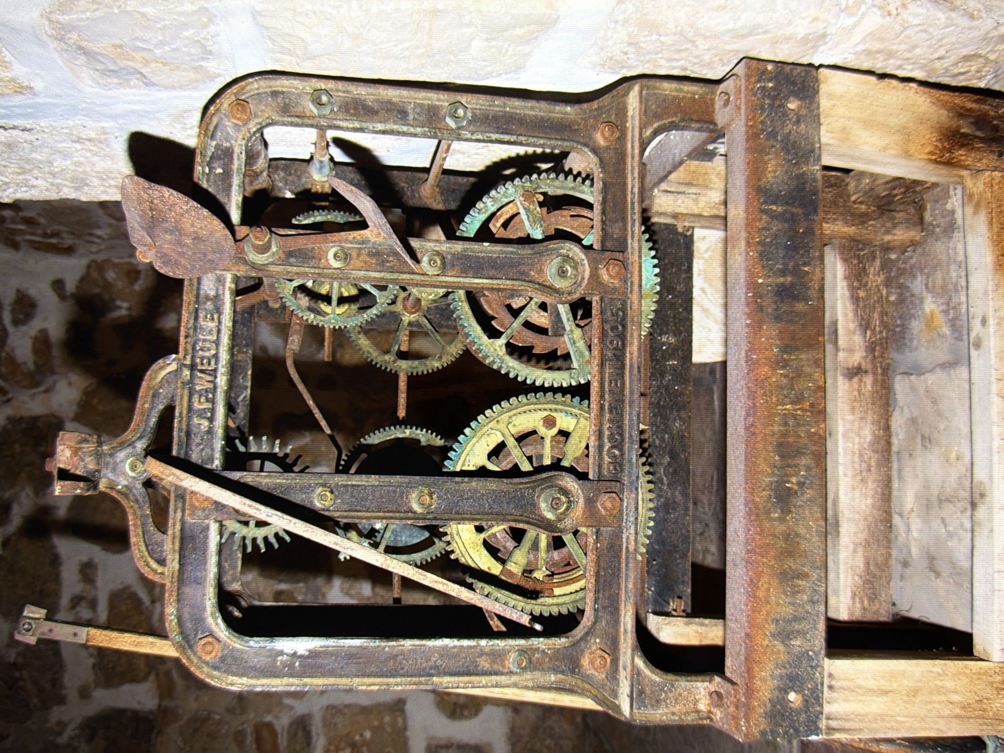 An antique mechanical device, possibly a printing press or clockwork mechanism, is set against a rough stone wall. The device is made of rusted metal with visible gears and levers, some of which are corroded and covered in green patina. A wooden frame supports the lower part of the machinery. The background consists of textured stone walls with variations in color and light. There are no people or animals visible in the image. The scene is devoid of any indication of location within the city of Kyiv, Ukraine.
