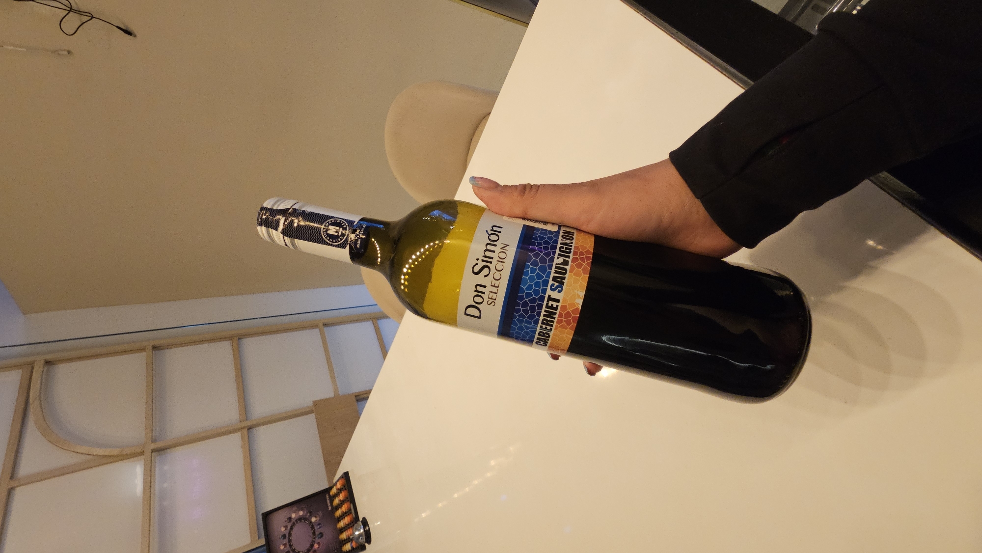 The image displays an indoor scene focused on a human hand holding a bottle of red wine. The hand, appearing female, has dark-painted fingernails and wears a black long-sleeved top featuring a thumbhole cuff on the wrist. It firmly grips a dark green glass bottle of "Don Simón SELECCION Cabernet Sauvignon" wine. The bottle label is multicolored, presenting "Don Simón SELECCION" in white text on a yellow field, above a mosaic-patterned band of blue and orange, and "CABERNET SAUVIGNON" in white text on a dark, patterned background. The bottle's neck is sealed with a blue and white striped foil that includes a white circular logo with an "M."
The bottle is held above a clean, reflective white tabletop. To the left on the table, a black box filled with multiple colorful Nespresso coffee capsules is open, accompanied by a small, metallic, bell-shaped object. The background on the left features a wall with a white panel adorned by a light-colored wooden lattice or grid structure. Further in the background, to the right, two light-colored, upholstered chairs with curved backs are partially visible. The wall behind these chairs is a plain light tan color, with black electrical cables loosely hanging in the upper right corner. The lighting appears to be bright and artificial, creating reflections on surfaces.
The environment suggests a commercial establishment such as a café, restaurant, or lounge, or a contemporary residential kitchen or dining area. The precise location within the city of Puebla, Mexico, is not discernible from the visual information provided in the image.