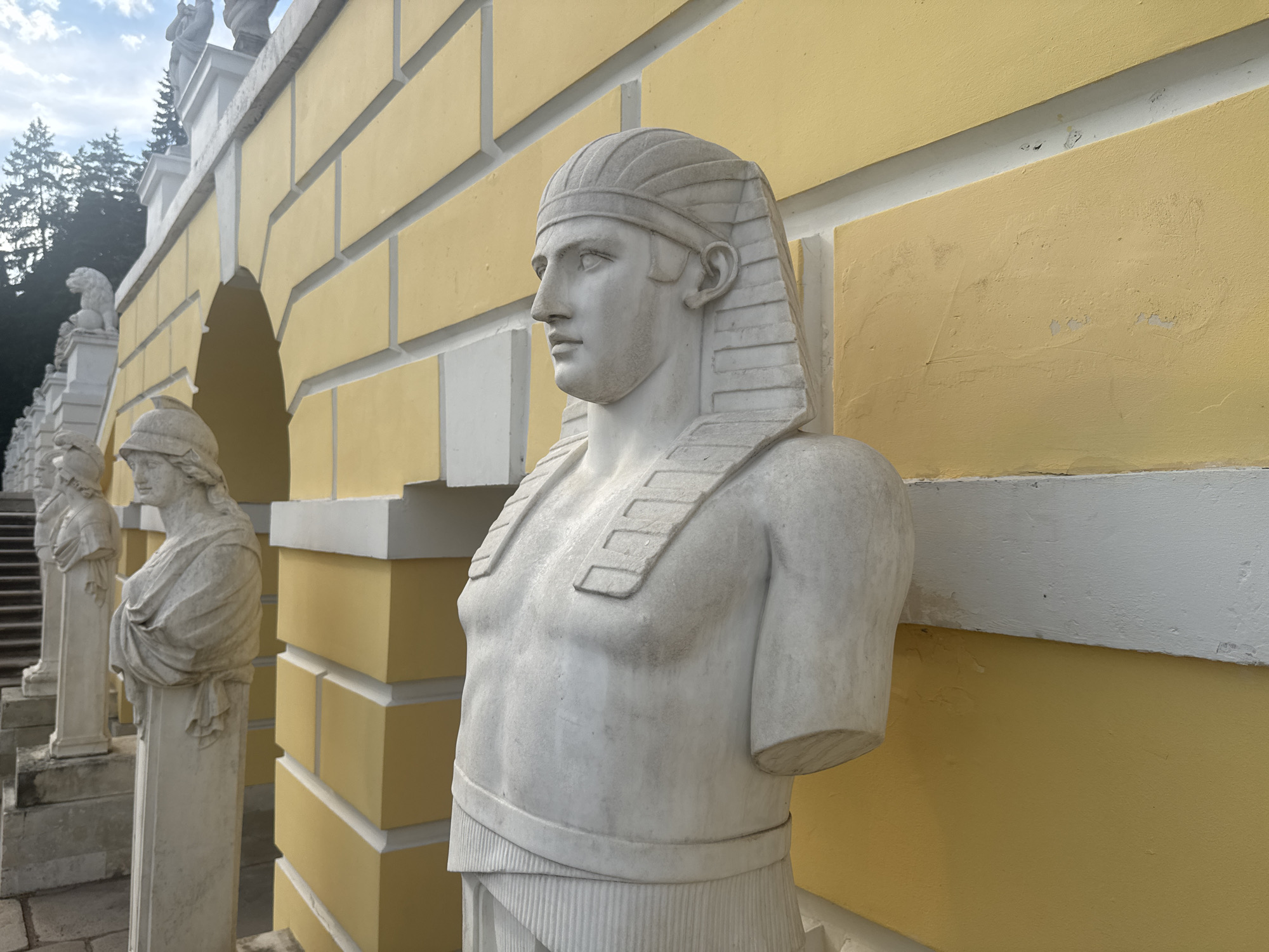 The image shows a sculpture of an Egyptian pharaoh in front of a yellow wall with white architectural details. The sculpture is made of white marble and depicts the upper body of a man wearing a nemes headdress and a pleated kilt. The pharaoh sculpture is positioned on the right side of the image, with its right shoulder and chest visible. To the left of the pharaoh sculpture, other marble sculptures are lined up, appearing as statues of people in classical attire. These statues are placed along a walkway or a balustrade, with steps visible behind them. The background features a yellow building with a white decorative border, and further left, trees and a cloudy sky are visible. The architecture suggests an outdoor setting, likely in a park or a historical estate. The location is identified as the estate "Arkhangelskoye" in the settlement of the same name, Russia, which is a well-known park and palace complex near Moscow.