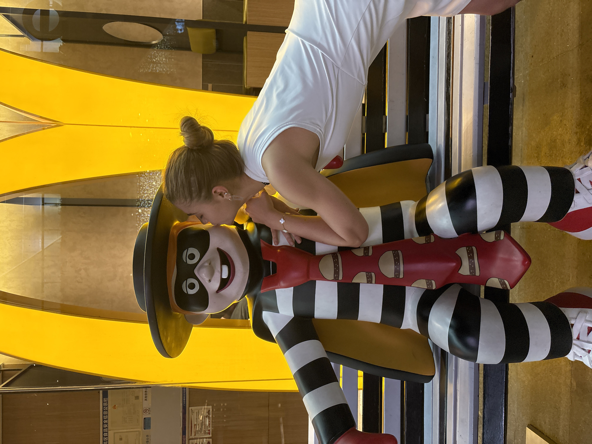 A woman in a white dress is leaning over and interacting with a statue of the McDonald's character Hamburglar. The Hamburglar statue is wearing his signature black and white striped outfit with a red tie adorned with hamburger images. He is also wearing a black hat and a black mask around his eyes. The background includes elements of a McDonald's restaurant interior, including a yellow arch-shaped feature. A sign containing legal notices is visible on the lower left. The location is in Panyu District, China.