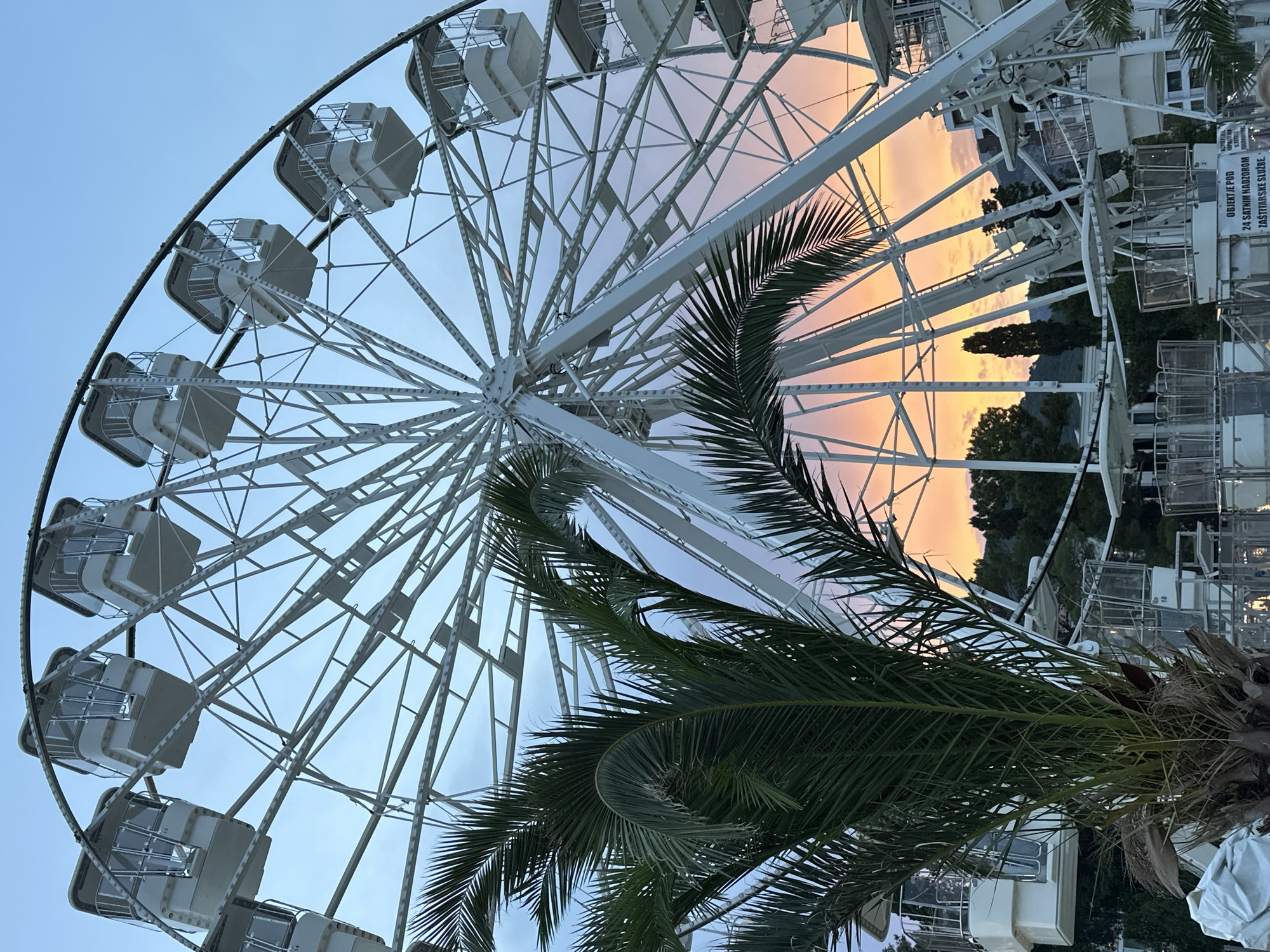 The image shows a Ferris wheel against a twilight sky in Budva, Montenegro. The Ferris wheel is white and has several gondolas. A palm tree with green fronds is in the foreground, partially obscuring the Ferris wheel. The sky exhibits a gradient of colors, transitioning from a pale blue at the top to warm hues of orange and pink near the horizon, indicating it is sunset. In the background, there are trees and buildings, suggesting an urban or coastal setting. The lighting and colors evoke a peaceful and serene atmosphere. Text is visible on a white panel attached to the Ferris wheel structure, which appears to be a warning sign. It reads "OBJEKT JE POD 24 SATNIM NADZOROM ZASTITARSKE SLUZBE". This translates from Croatian to "THE OBJECT IS UNDER 24-HOUR SURVEILLANCE BY SECURITY SERVICE".