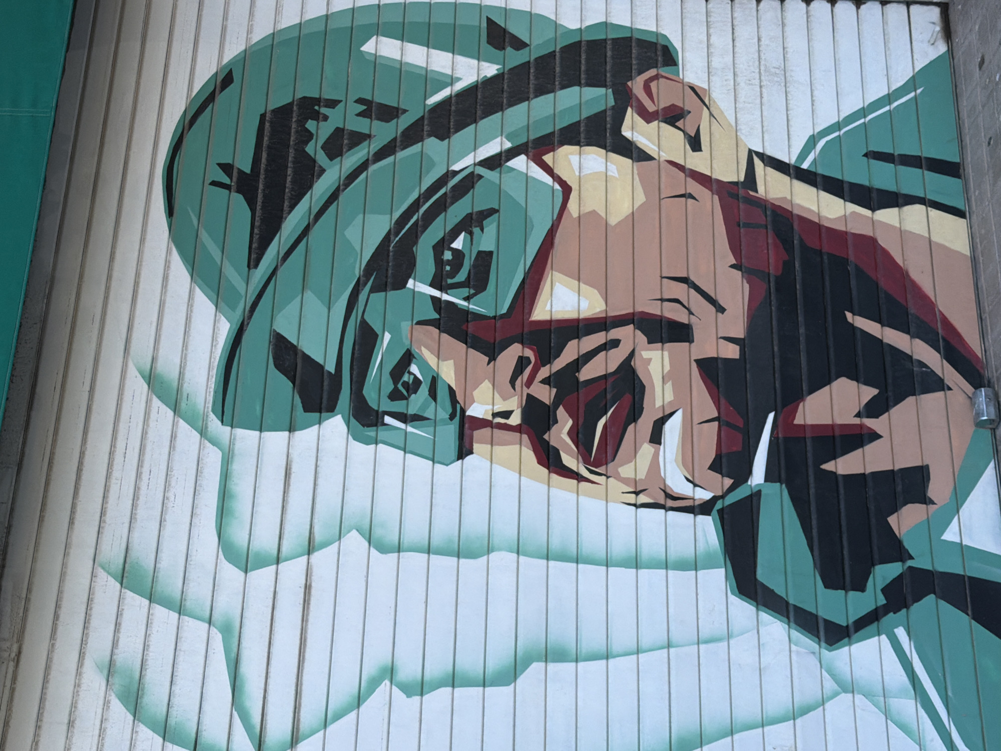 A stylized, graphic mural is painted on a corrugated metal roll-up door in a city. The mural depicts a close-up of a person's face, rendered in a bold, angular style with a limited color palette. The person wears a green helmet and goggles, with a green and black collar visible below the chin. Their skin tones are depicted in shades of tan and red, with sharp, geometric lines. Behind the face, faint, translucent outlines of what appear to be abstract shapes or figures in a light blue-green hue are visible, creating a layered effect. The metal door itself has horizontal grooves, which are partially obscured by the paint. The lower edge of the door rests on a gray concrete surface, and a small, cylindrical metal object, possibly a lock or stopper, is visible at the bottom right corner. The overall impression is one of a graphic, possibly political or propaganda-style artwork.