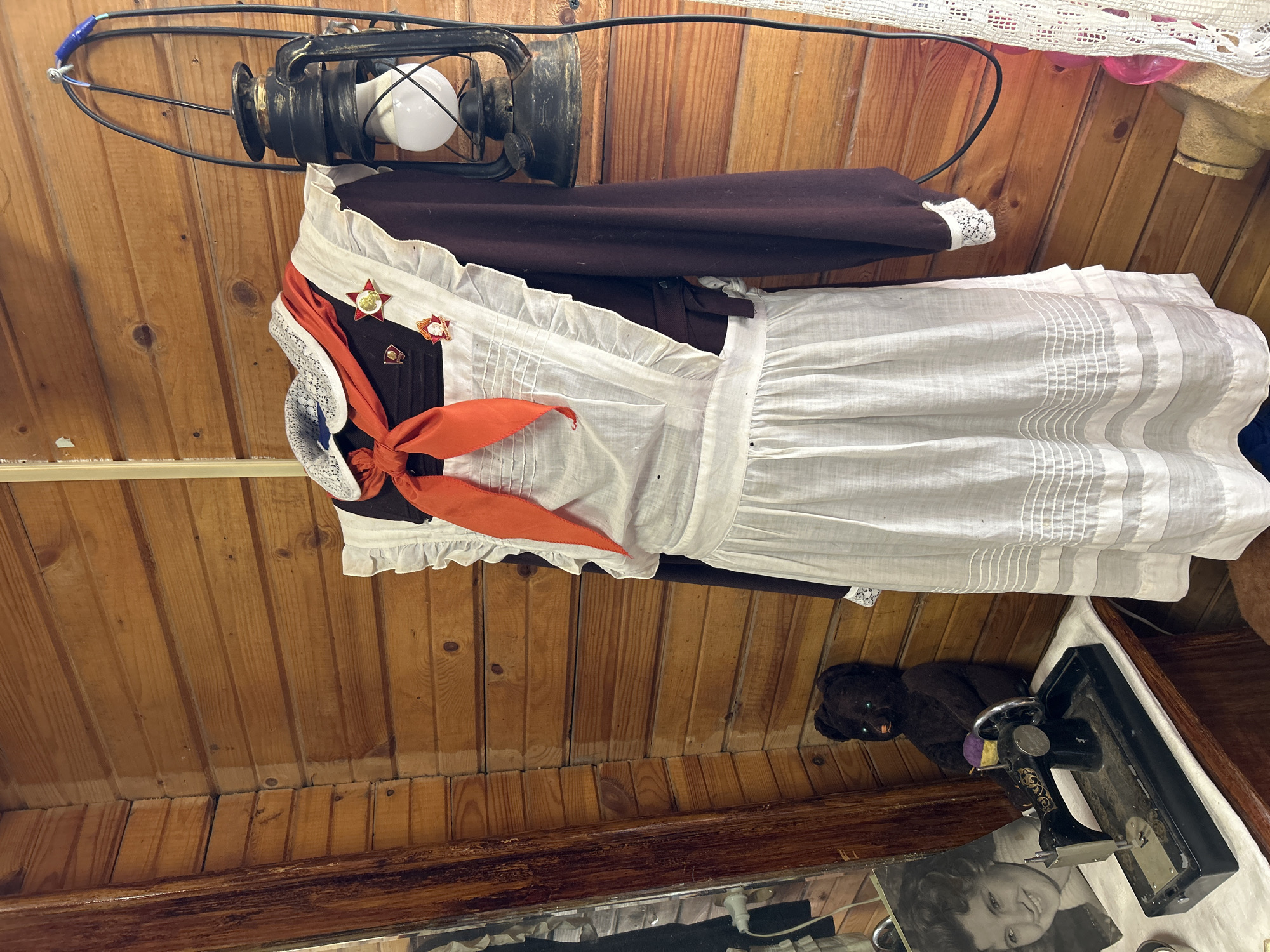 The image shows a room with a wooden ceiling. Hanging from the ceiling is a vintage-style lantern. A Soviet-era school uniform is also hanging in the room. The uniform consists of a brown dress with white lace trim, a white apron, and an orange pioneer scarf. There are also Soviet-era pins attached to the dress. On a table below is an antique sewing machine and a stuffed bear. Also on the table is a black and white photograph of a woman. The room appears to be part of a museum exhibit. While the location isn't definitively confirmed as Moscow, Russia, the presence of the Soviet school uniform is a strong indicator.