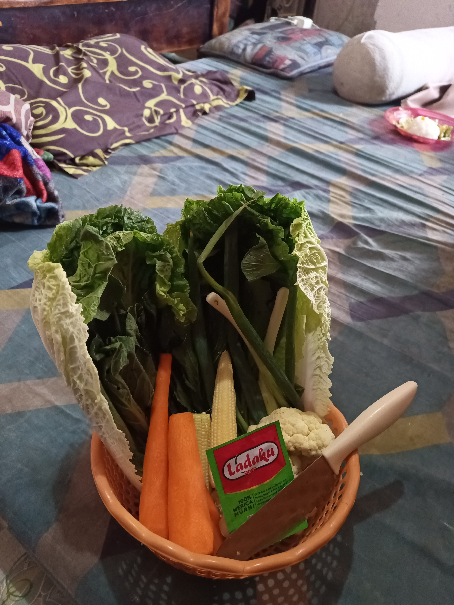 The image shows a basket of fresh vegetables placed on a bed with a patterned sheet. The vegetables include Napa cabbage, carrots, baby corn, spring onions, and cauliflower. A packet of "Ladaku" seasoning and a knife with a light-colored handle are also in the basket. The background reveals a wooden headboard and pillows on the bed, suggesting an indoor setting. The patterned bedsheet and the general ambiance do not provide any specific clues to pinpoint the location to a particular area within South Jakarta, Indonesia.