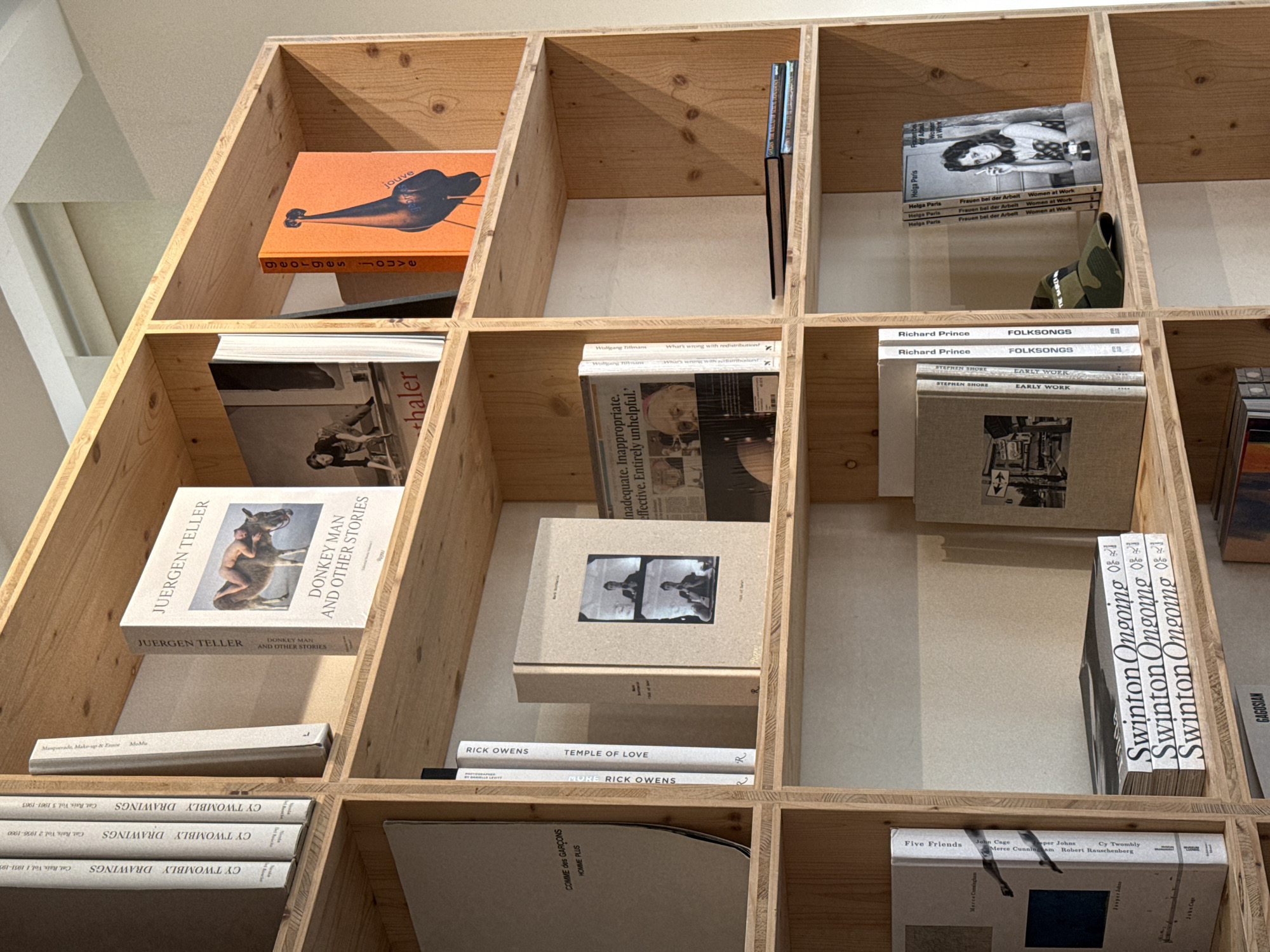 The image shows a wooden bookshelf with several books and items arranged in its compartments. The books cover a range of subjects, including art, photography, and fashion. Some of the notable titles and authors include "Donkey Man and Other Stories" by Juergen Teller, "CY TWOMBLY DRAWINGS," "TEMPLE OF LOVE" by Rick Owens, "Frauen bei der Arbeit Women at Work" by Helga Paris, "FOLKSONGS" by Richard Prince, and "EARLY WORK" by Stephen Shore.
There is a camo hat, as well, in the shelf.