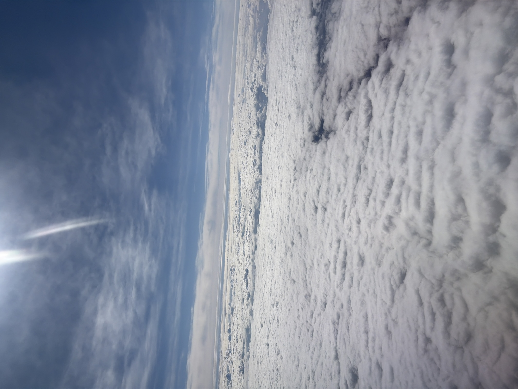 The image shows an aerial view of clouds and sky. A bright sun is visible on the left, with the sky transitioning to a deep blue further up. A dense layer of white, textured clouds fills the right side of the frame. A strip of thinner clouds or haze separates the sky from the dense cloud layer. There is no indication of the location being Sucha Beskidzka, Poland.