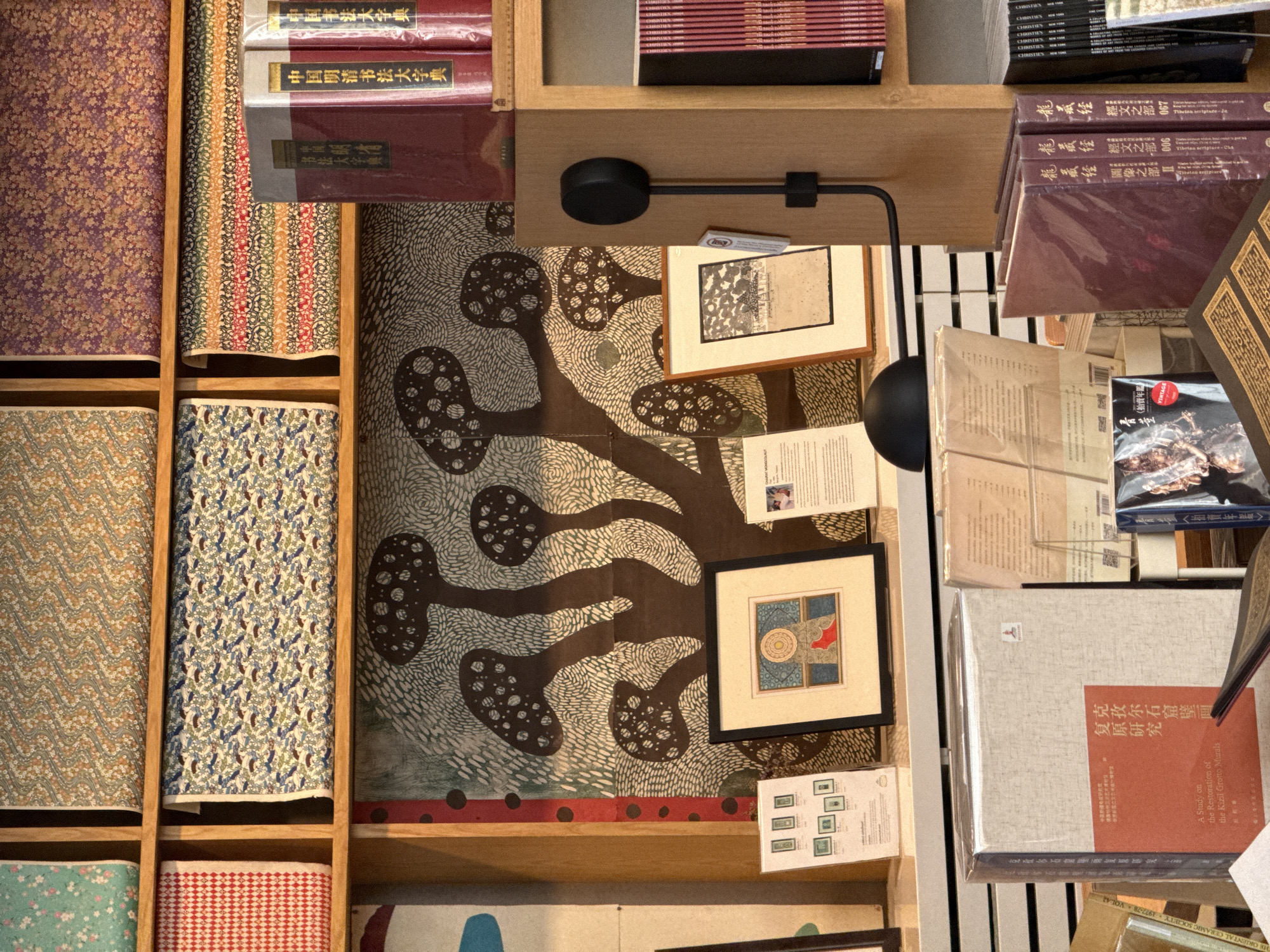 The image shows a well-lit interior space, possibly a shop or library, in Bangkok, Thailand, due to the presence of books with Chinese characters and traditional Thai patterns. A wooden shelving unit dominates the scene, displaying a variety of items including books, patterned textiles, framed artworks, and paper documents. The textiles showcase floral and geometric designs, while the books range from modern publications to older volumes with worn covers. A modern black lamp is mounted on the shelf, providing direct light. The framed artworks include drawings and illustrations, while loose papers appear to be documents or notes. The books on the right have Chinese script. The composition suggests a curated display of art, literature, and historical artifacts.