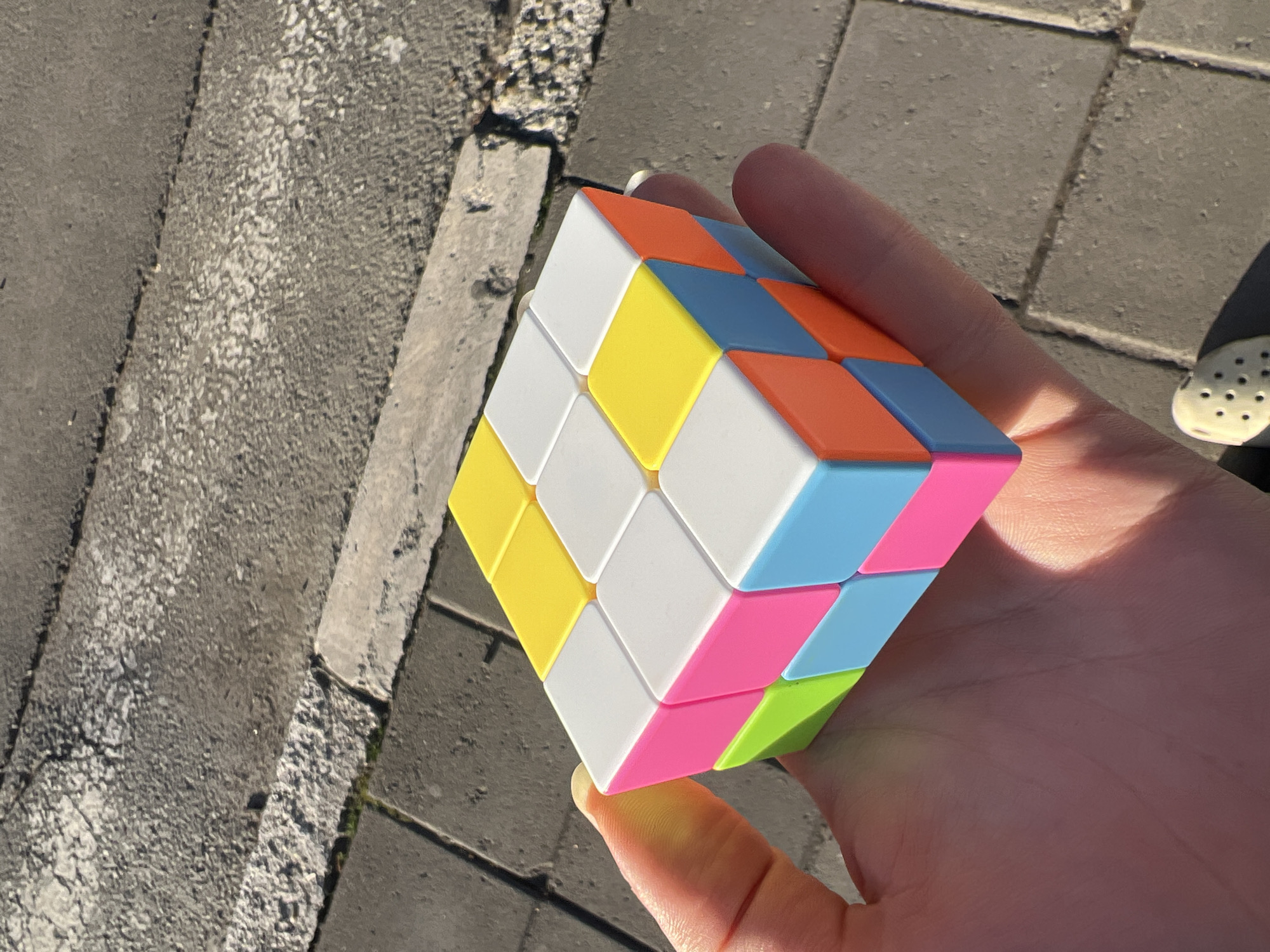 A person's hand holds a Rubik's Cube. The cube's visible sides display yellow, white, orange, blue, pink, and green colors. The background shows a concrete pavement and a curb. A partial view of a shoe, possibly a sandal, is visible on the right. The location is urban, potentially Belgrade, Serbia, but there are no definitive landmarks or contextual clues to confirm the city.