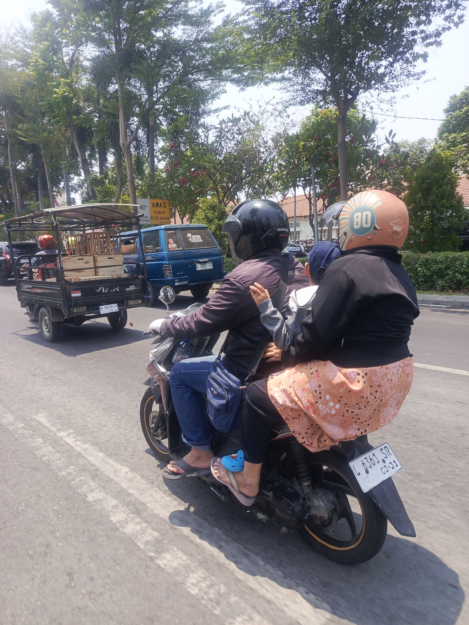In a street scene in Surabaya, Indonesia, three individuals ride a motorcycle. A male driver in a dark jacket and blue jeans, wearing a black helmet, operates the vehicle. Seated behind him is a woman wearing a black jacket and an orange floral skirt, with a blue child's hat on her head. A small child, likely the one wearing the blue hat, is positioned between them, with their arm around the woman. The child is wearing a grey long-sleeved shirt. The motorcycle has a license plate that reads "L 6361 SR" with the date "03-21." In the background, a blue pickup truck is visible, carrying a load of wooden crates. A smaller black truck with a canopy is to the left of the pickup, also carrying cargo. The environment is characterized by trees lining the street and buildings in the distance, suggesting an urban setting. The weather appears sunny.