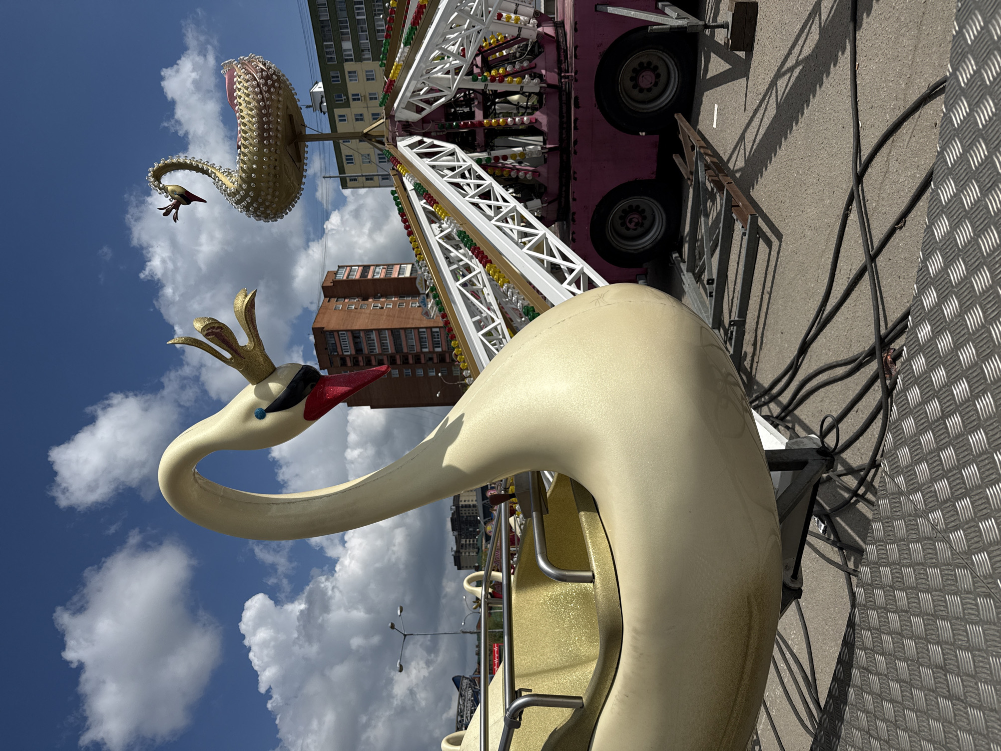 A low-angle shot of a carousel with swan-shaped seats in Dmitrov, Russia. The swan seat in the foreground is painted a light yellowish color and has a decorative crown on its head. Other swans and ride equipment are visible. The background shows a blue sky with puffy white clouds and buildings typical of an urban area. The pavement appears to be metal. Electrical cables lie nearby.