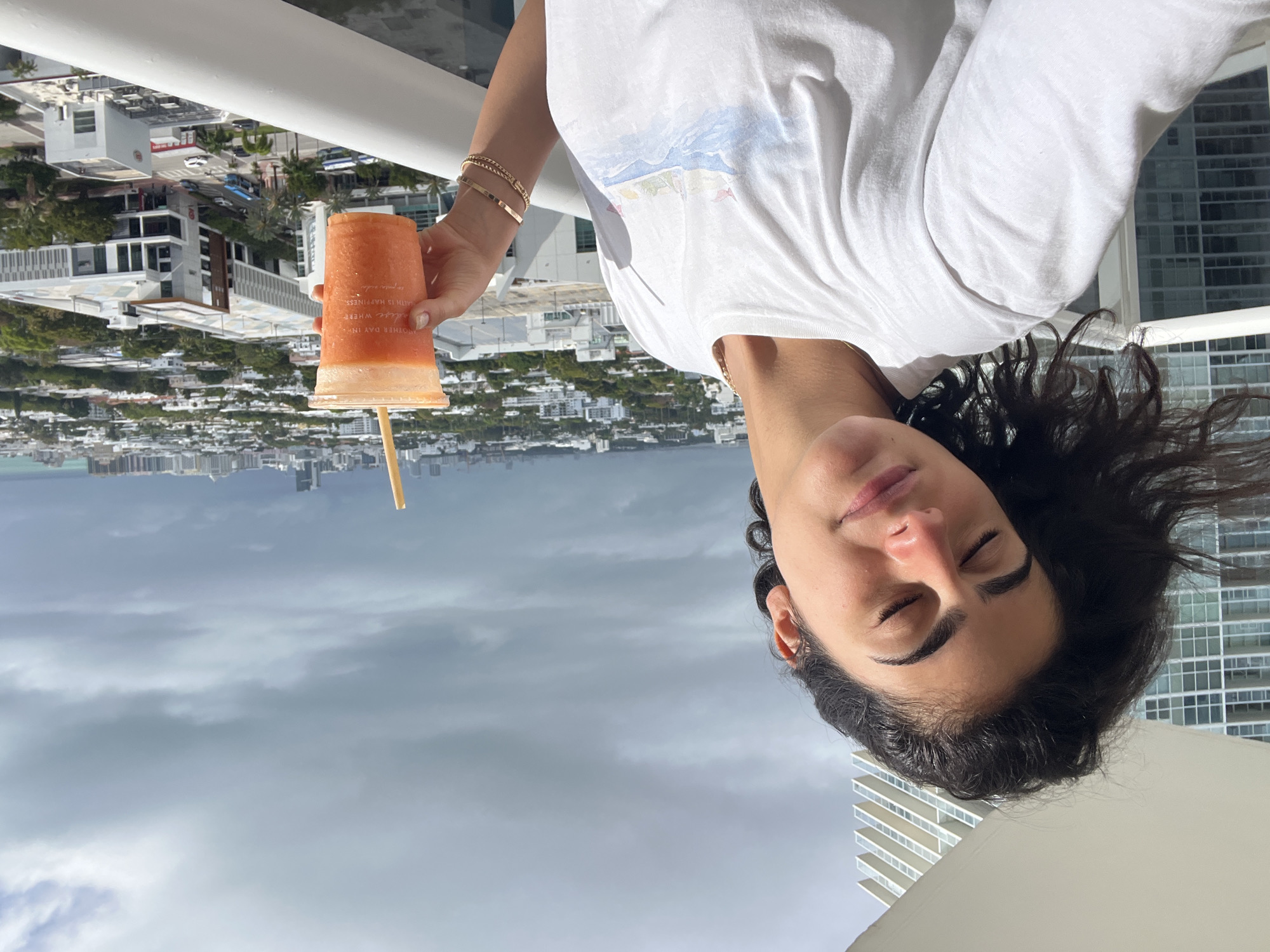 A medium-level shot depicts a woman holding a drink on a balcony in Miami Beach, United States. The woman is wearing a white t-shirt and has dark hair. She is holding a tall, transparent plastic cup containing an orange beverage with a straw. Her eyes are closed. She wears two gold bracelets on her left wrist.
In the background, there is a cityscape, including buildings and foliage. The sky is overcast with grey clouds. White balcony railings are visible.
Based on the architectural style and general environment, the location is identifiable as Miami Beach, United States.