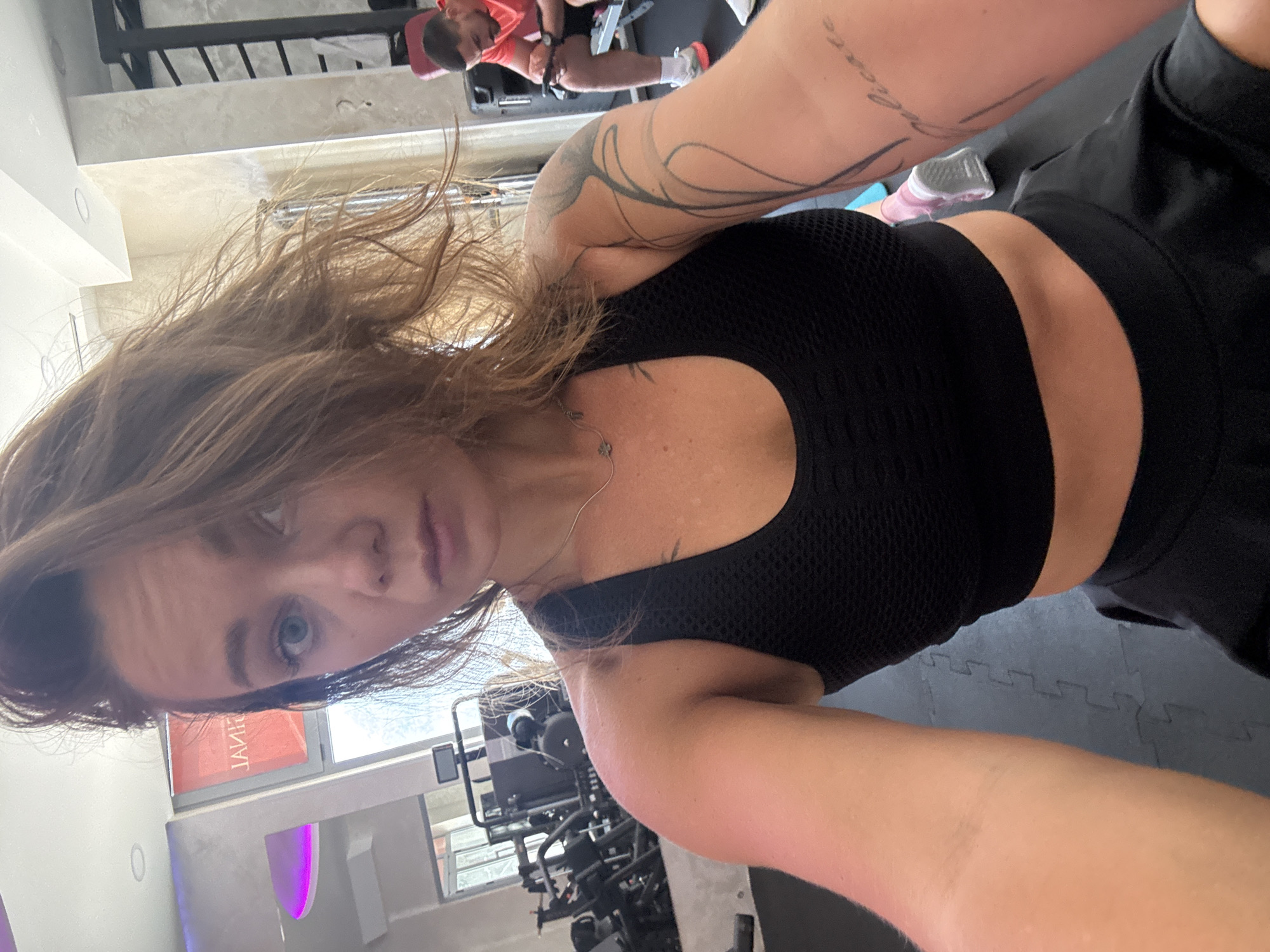 This is a selfie taken in a gym in Budva, Montenegro. A woman with long, wavy brown hair is in the foreground, wearing a black sports bra and black shorts. She has tattoos on her arms. In the background, a man wearing a red shirt is visible, sitting on a weight machine. The gym appears to be modern, with various fitness equipment and padded flooring. The lighting suggests it could be daytime. The woman's expression is neutral. A sign with Cyrillic text is partially visible, reading "ЈАНИЦ".