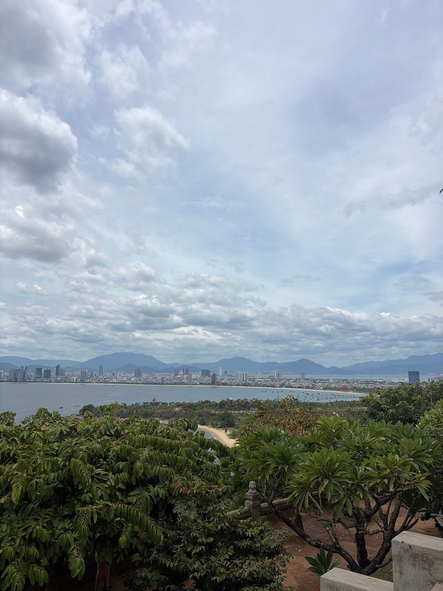 This image captures a panoramic view of the city of Đà Nẵng, Vietnam, from a higher vantage point. In the foreground, lush green trees with broad leaves dominate the scene, with a hint of a stone structure on the right side suggesting a man-made element in the natural landscape.
Beyond the trees, a sandy beach curves along the coastline, leading the eye to the expansive blue ocean. The water is dotted with numerous small boats, indicative of an active harbor or fishing community.
In the midground, the city of Đà Nẵng unfolds with a dense collection of buildings, including high-rise structures that define its urban character. A long stretch of buildings runs parallel to the coastline, suggesting a developed waterfront. A prominent, modern skyscraper with a crane nearby stands out on the right side of the city skyline.
The background is dominated by a range of hazy mountains, their peaks softened by the distance and atmospheric conditions. The sky above is a canvas of dramatic clouds, with patches of lighter blue sky interspersed with heavy, grey formations, hinting at a dynamic weather pattern. The overall impression is one of a vibrant coastal city nestled against a backdrop of natural beauty.