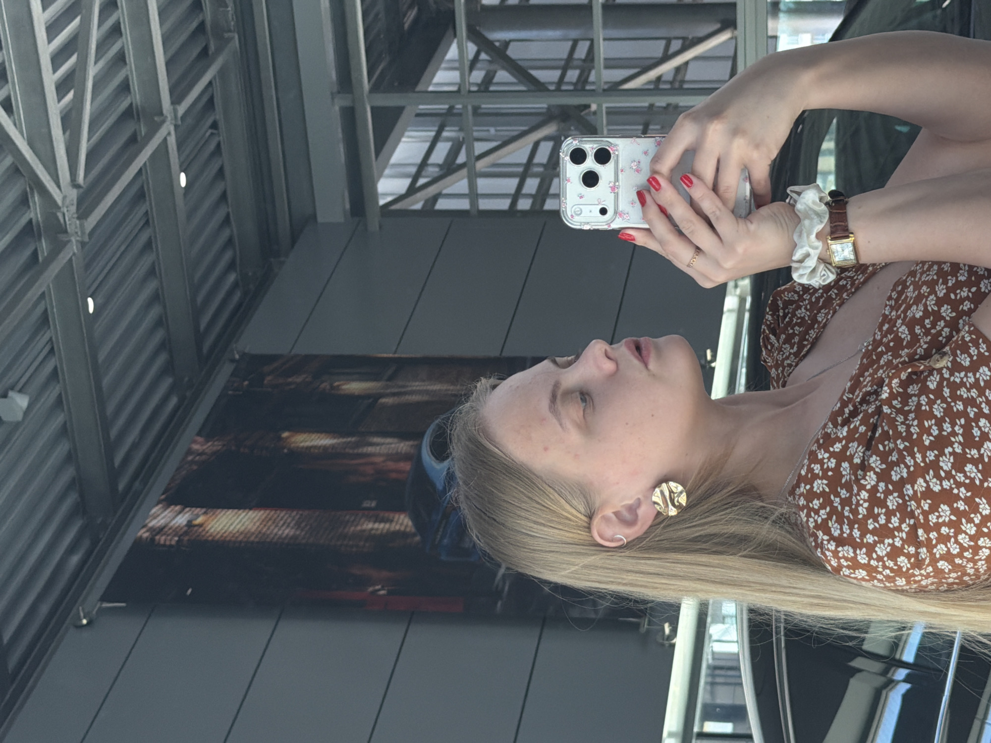 A woman is holding a smartphone with a floral case in her left hand, and the fingers of her right hand are on the screen, while she takes a selfie. She has long blonde hair, a gold earring, a scrunchie around her right wrist, and a brown floral print top. A wristwatch is on her right wrist. The background suggests a modern building with metal beams and panels and a large glass surface. It is in the city of Izhevsk, Russia.