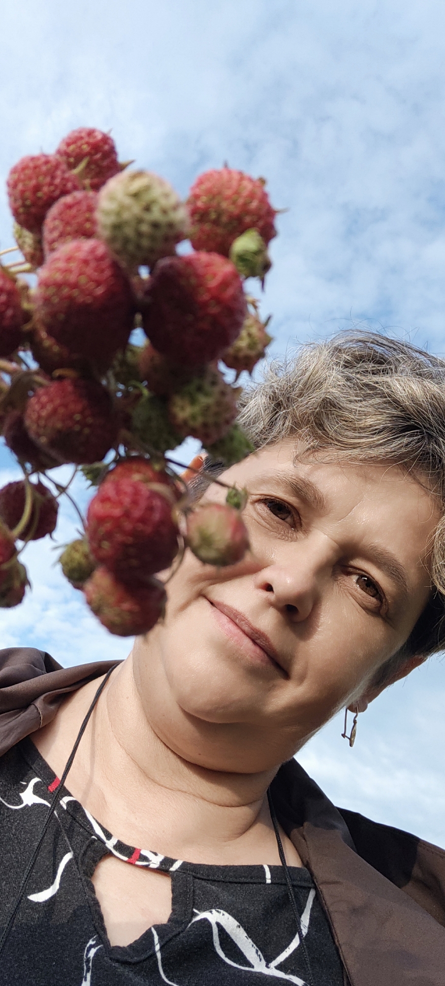 A close-up, low-angle shot captures a woman's face from the chest up, with a cluster of red and green berries in the foreground obscuring part of her face. The woman has short, graying hair with streaks of blonde and appears to be looking upwards. Her eyes are brown, and she has a gentle smile. She is wearing a dark brown jacket over a black and white patterned top with a cutout at the neckline. A thin black cord necklace is visible. She has a small, delicate earring in her left ear. The background is a bright blue sky with scattered white clouds, suggesting daytime and fair weather. The berries, some ripe red and others unripe green, are hanging from a branch, possibly from a raspberry or strawberry plant, as suggested by the location context of Bogdashkino, Russia. The overall mood is relaxed and natural, possibly indicating a moment of enjoying nature or harvesting fruit. There is no visible text in the image.