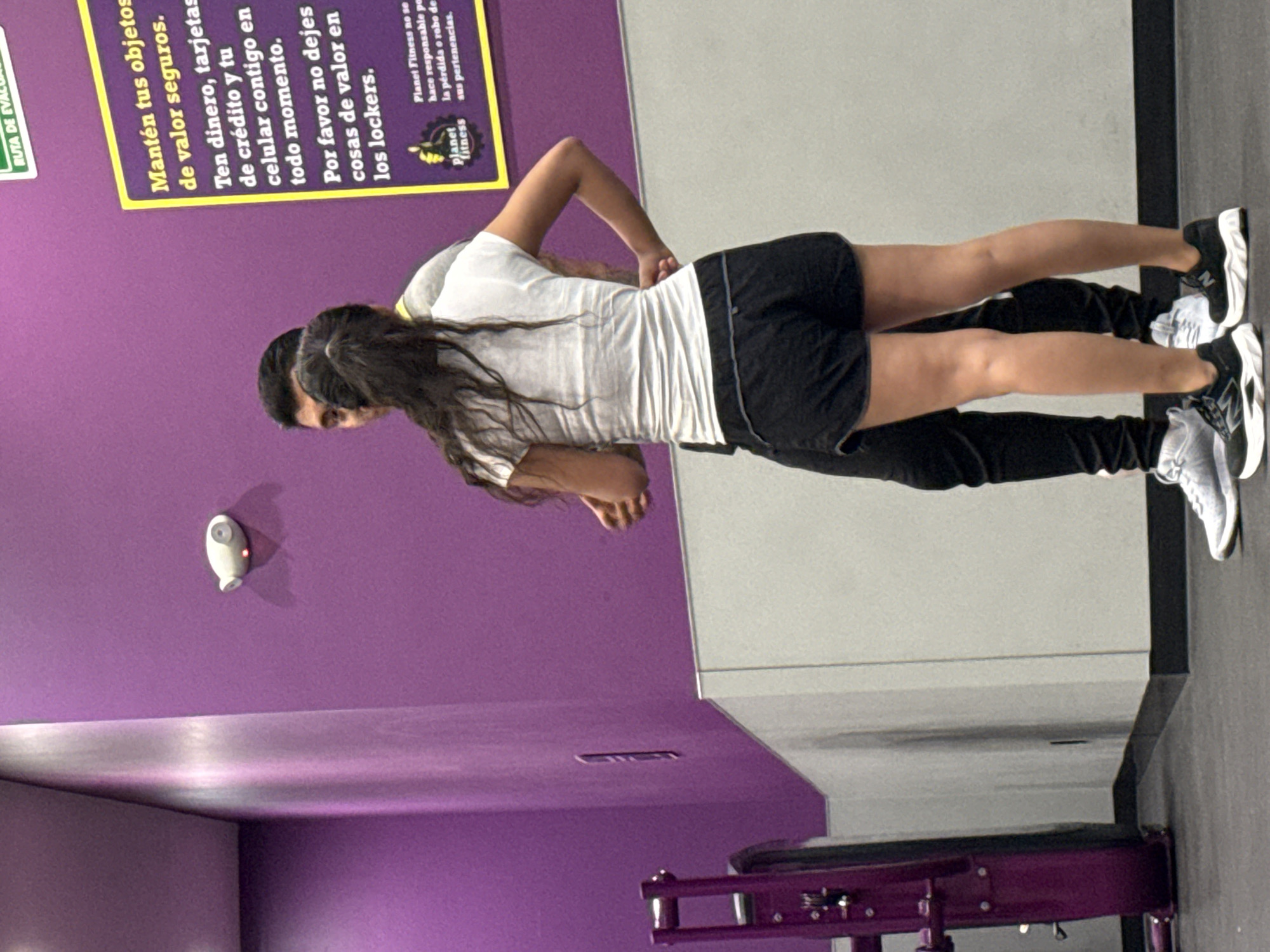 Two individuals are captured in an indoor gym setting, characteristic of Planet Fitness, with a prominent purple wall in the background. The image is taken at a slight upward angle, showing the gym's vibrant purple walls, a light grey wall or mirror, and a dark grey floor. Part of a purple gym machine is visible in the lower right corner.
In the foreground, a woman with long, dark hair, wearing a white t-shirt, black shorts, and black and white New Balance sneakers, is positioned behind a man. Her hands are placed on his lower back or waist, suggesting a partner exercise or stretch. The man, visible from the back, is wearing dark pants and white and black New Balance sneakers. Both individuals appear to be actively engaged in a fitness activity.
On the purple wall to the left, a white sensor or camera with a red indicator light is mounted. Above them, a large yellow sign with Spanish text is prominently displayed. The text on the sign advises patrons: "Mantén tus objetos de valor seguros. Ten dinero, tarjetas de crédito y tu celular contigo en todo momento. Por favor no dejes cosas de valor en los lockers." (Keep your valuable objects safe. Keep money, credit cards, and your cell phone with you at all times. Please do not leave valuable things in the lockers.) The sign also includes the Planet Fitness logo and states, "Planet Fitness no se hace responsable por la pérdida o robo de sus pertenencias." (Planet Fitness is not responsible for the loss or theft of your belongings.) A partial green sign with "RUTA DE EV" (likely "RUTA DE EVACUACIÓN" - evacuation route) is also visible. The scene is likely taking place in Puebla, Mexico, given the Spanish text and the context of the location.