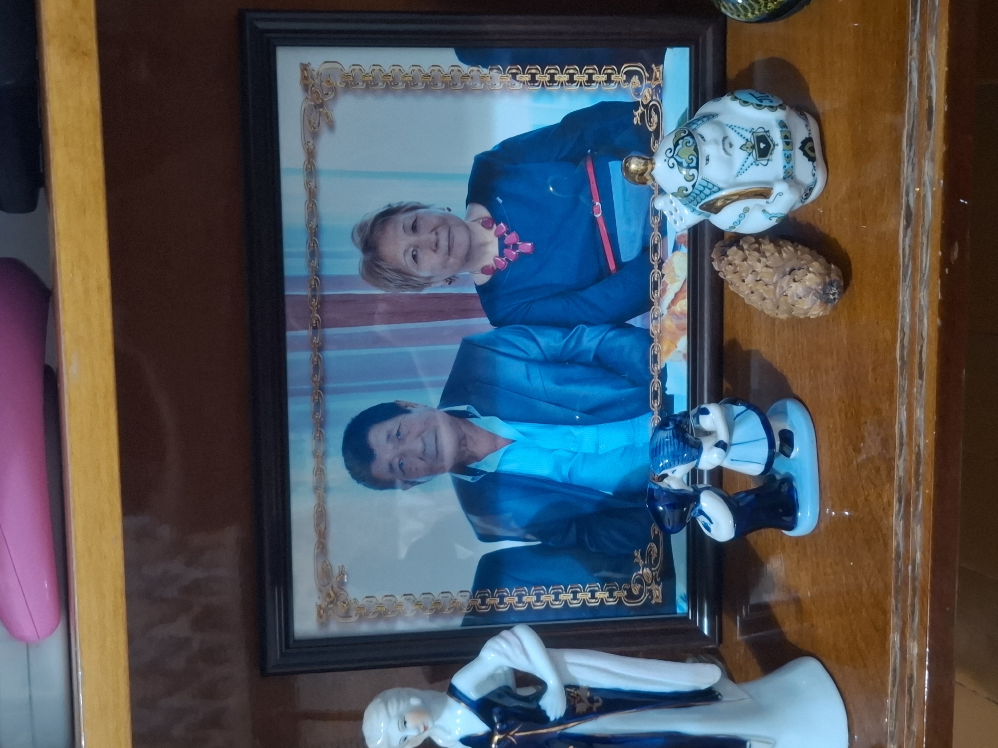 The image depicts a framed photograph of an elderly Asian couple, likely husband and wife, displayed on a wooden shelf. The man, on the left, wears a blue shirt and a dark suit jacket. The woman, on the right, is adorned with a large, pink, statement necklace and a dark blue dress with a red belt. The photograph has an ornate gold border. To the left of the photograph is a tall, slender porcelain figurine of a woman in a dark blue and white dress. In front of the photograph, there are several decorative items: a small ceramic figurine of two children, a pinecone, and a decorative white teapot with blue and green accents, resembling a traditional warrior or deity. The shelf appears to be in a home environment, with the background suggesting a wall with some decorative pattern or wallpaper. The overall impression is a personal display of cherished memories and decorative objects. The image is too general to pinpoint a specific location within Almaty, Kazakhstan.