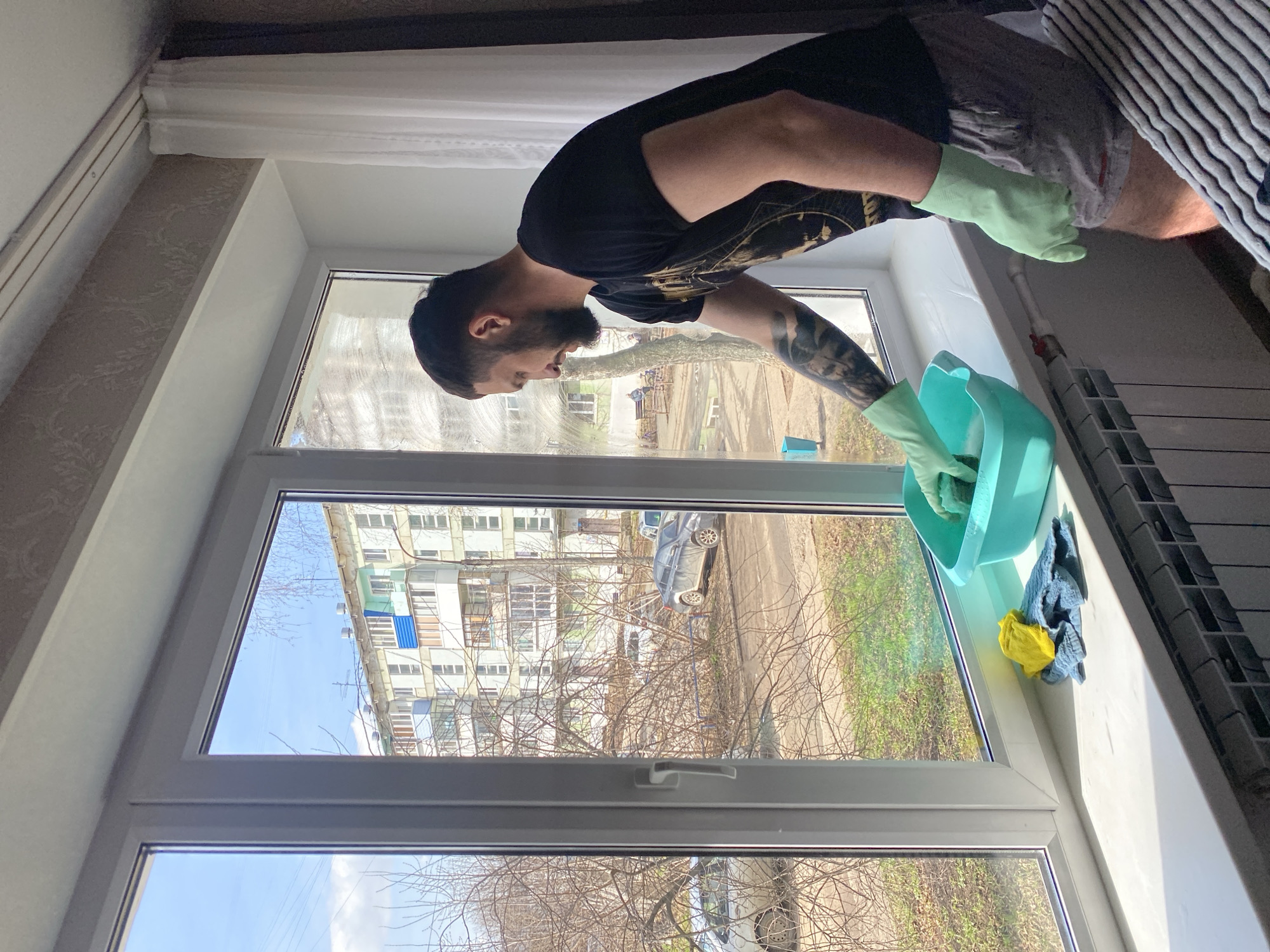 A man is cleaning a window in a building located in Chaikovsky, Russia. The man is wearing a black t-shirt, grey shorts, and green gloves. He is holding a green bucket and a sponge, and he is wiping the window with the sponge. Outside the window, there are several buildings, trees, a parked vehicle, and a grassy area. The sky is blue. Inside the building, there is a radiator next to the window. There is also a patterned wallpaper on the wall. The scene appears to be taking place during the day.